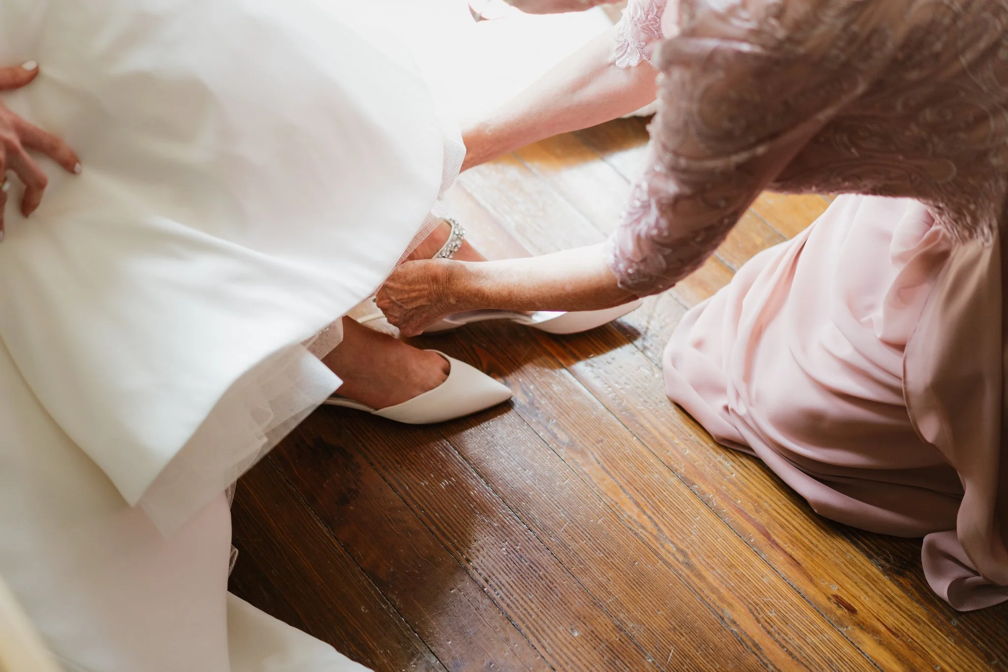 A person in a white dress, possibly a wedding gown, holding hands with another individual wearing a pink dress with lace detail, both sitting on a wooden floor.