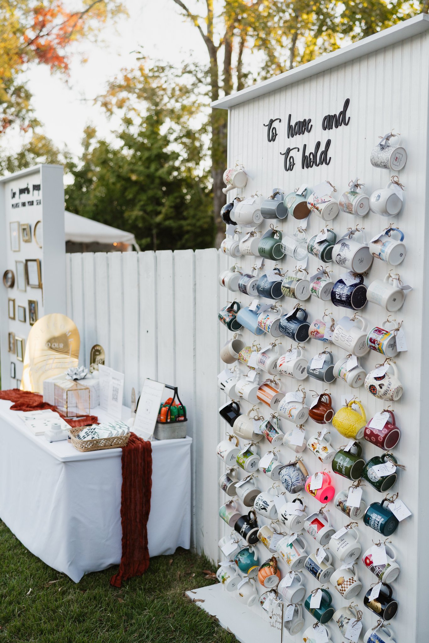 Outdoor craft or thrift fair with a white display board filled with various decorated and plain mugs hanging on hooks. A white table with items and a red table runner stands next to the display, with a basket of small objects on top. There are trees 