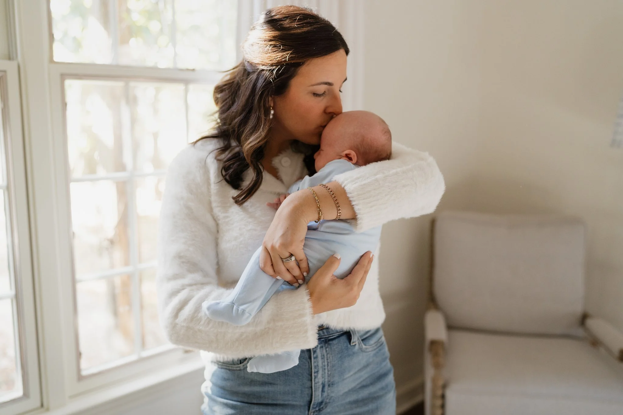 A woman with wavy brown hair, wearing a white sweater and jewelry, holding and kissing a sleeping baby in Memphis, TN. 