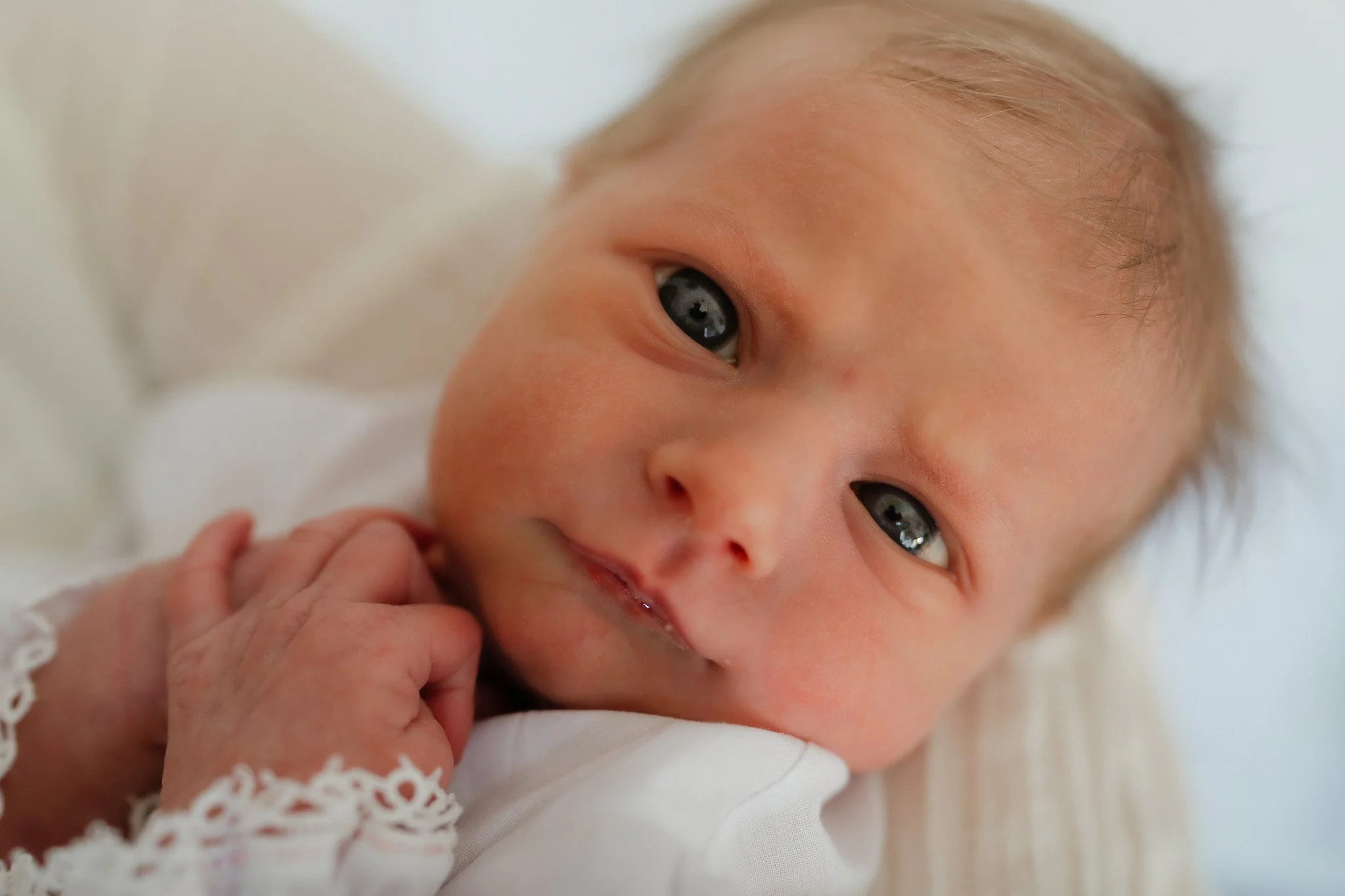 Close-up of a baby's face with blue eyes, light skin, and blonde hair, lying on a white surface, gently holding a tiny finger in Memphis, TN. 