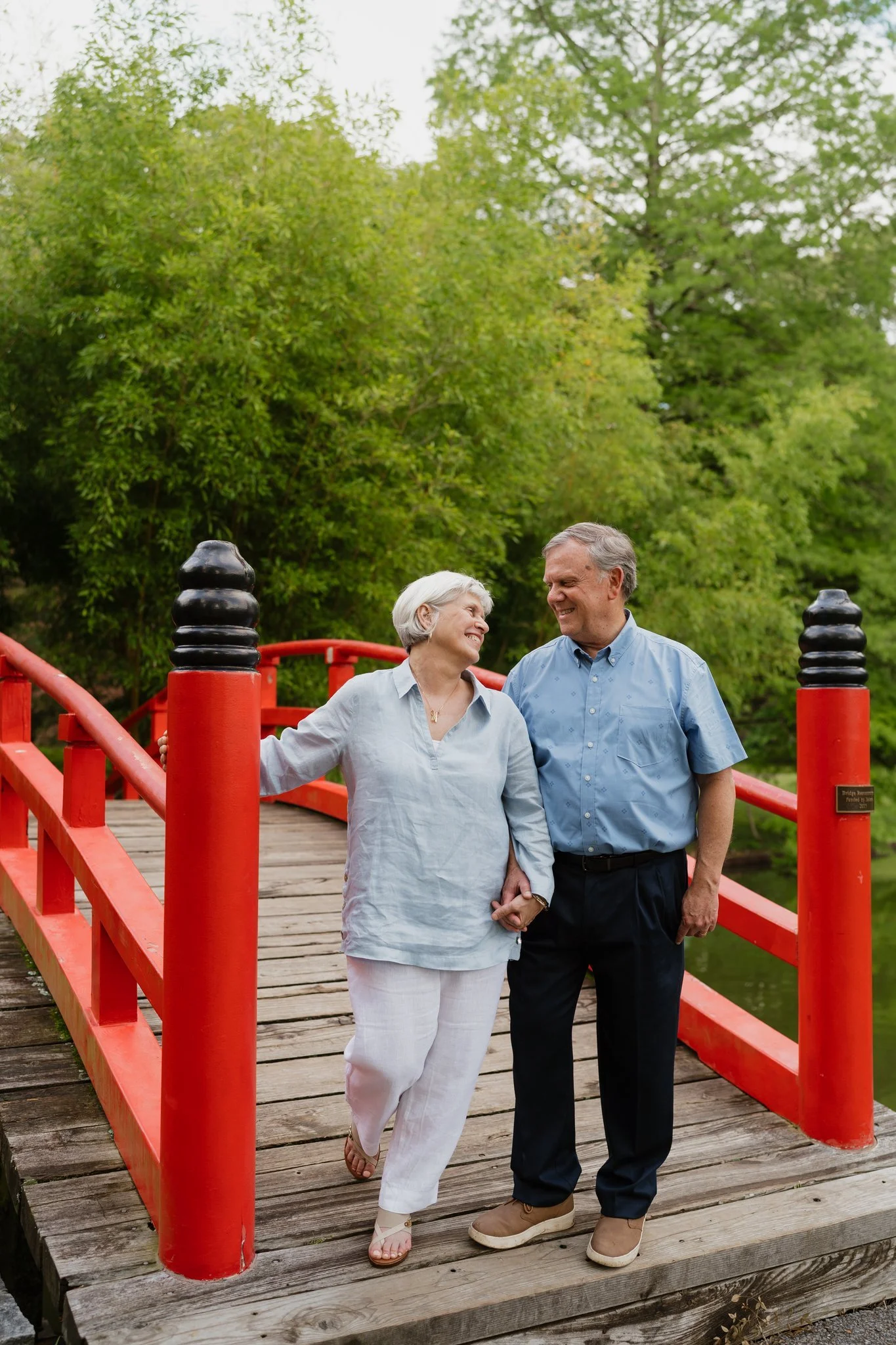An elderly couple, a woman in white pants and a man in dark pants, holding hands and smiling at each other on a wooden bridge with red railings, surrounded by green trees in Memphis, TN. 