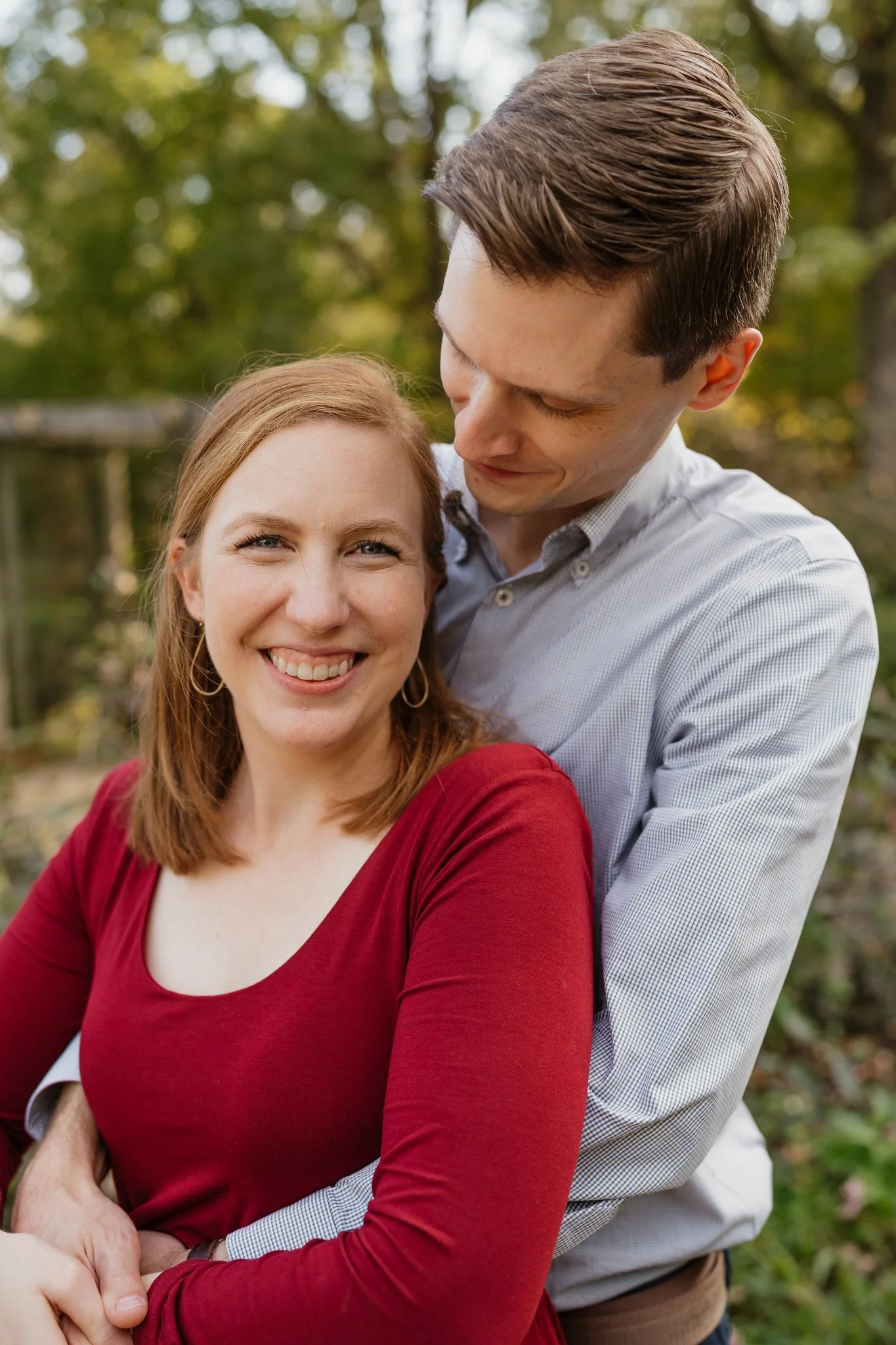 A smiling woman with reddish-brown hair wearing a red long-sleeve top is being embraced by a man with short brown hair, wearing a light blue checkered shirt, outdoors with trees in the background in Memphis, TN. 