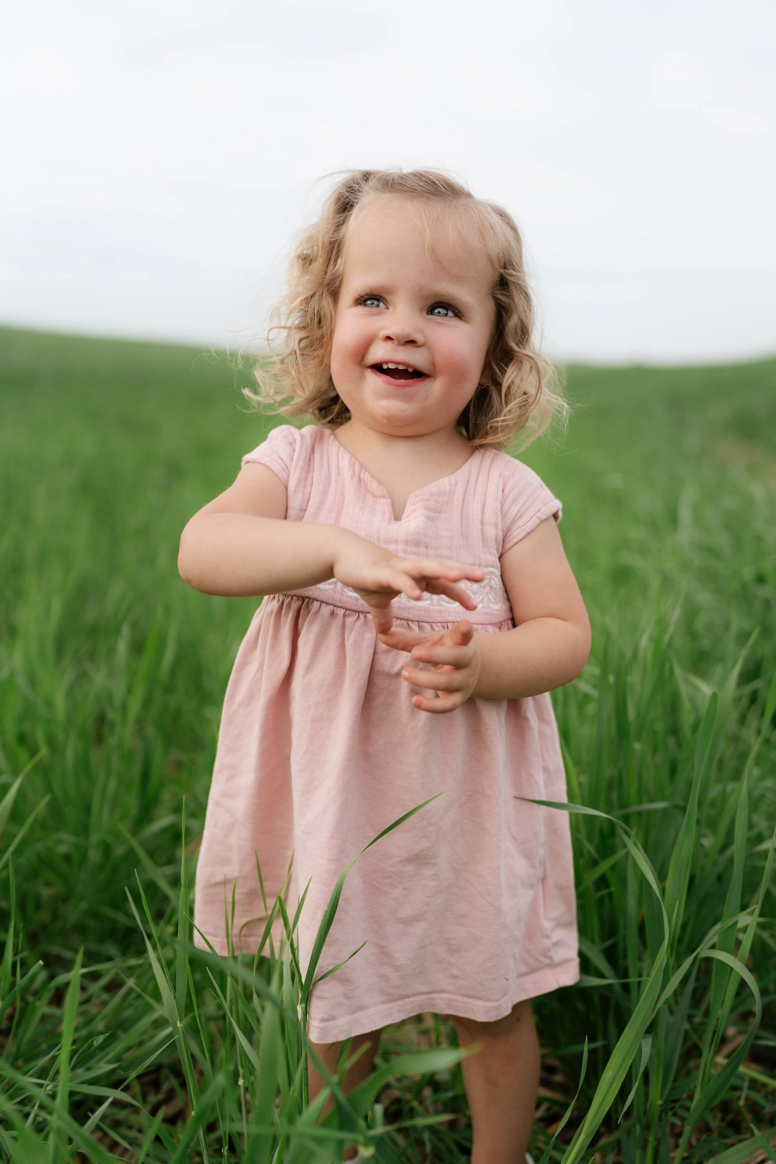 A young girl with curly blonde hair and blue eyes smiling and playing in a grassy field, wearing a pink dress in Memphis, TN. 