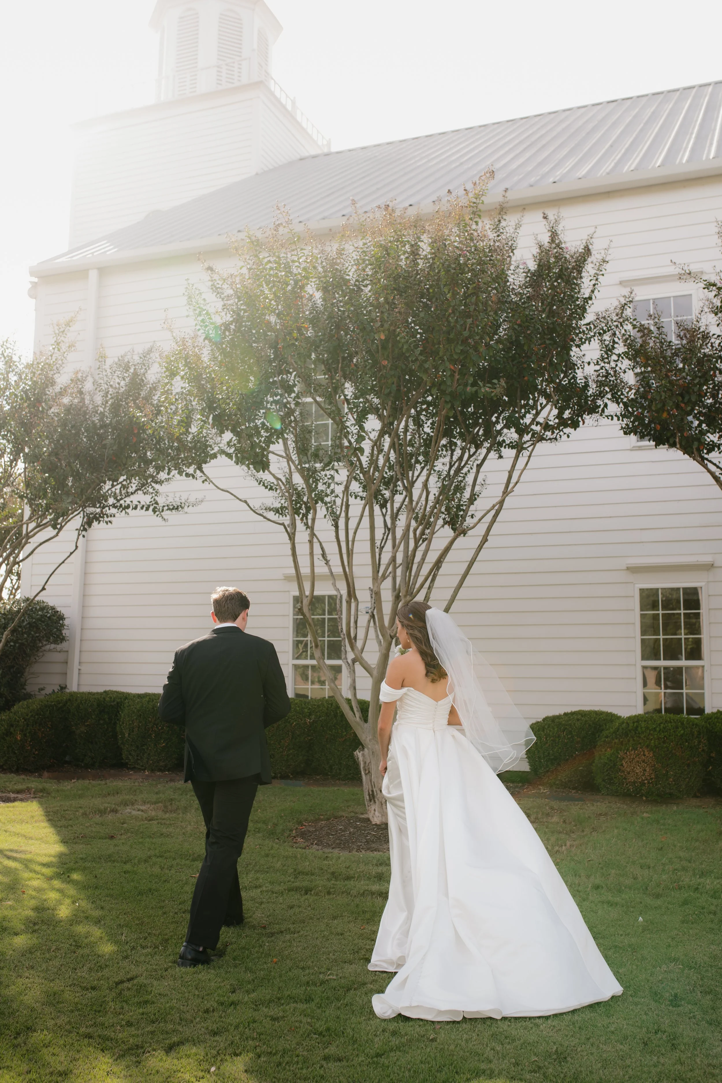 A bride and groom standing outside on a lawn in front of a white house, with trees and bushes around them, during daytime, with sunlight creating a flare in Memphis, TN. 