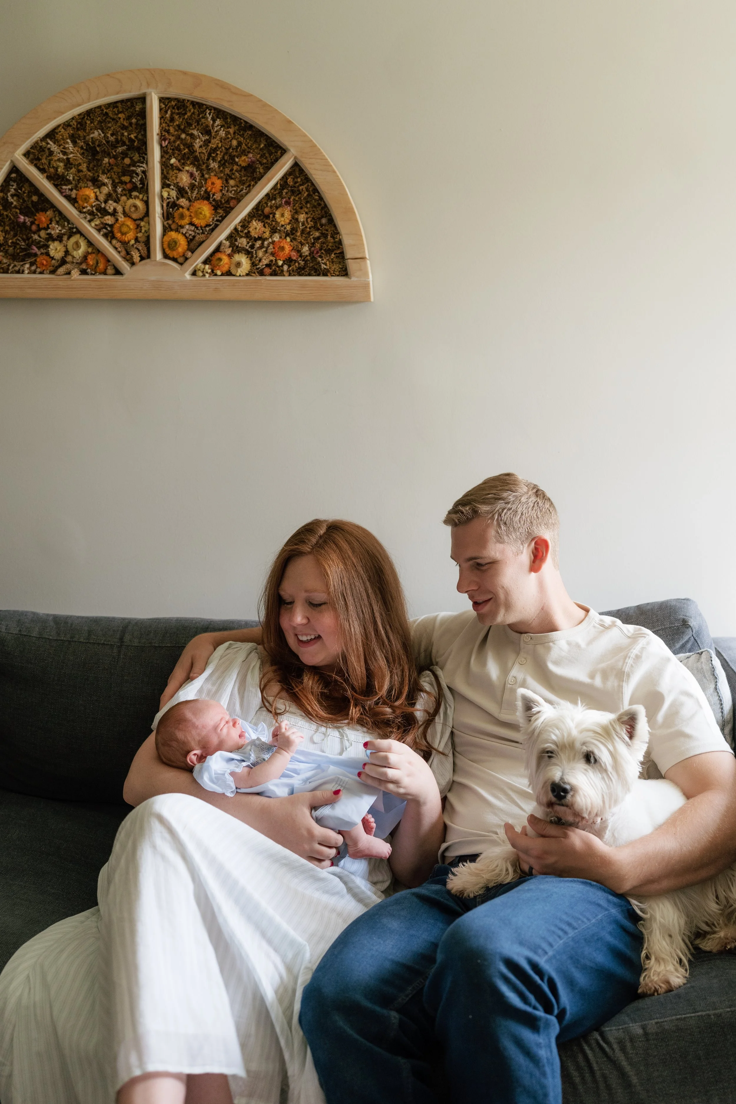 A family sitting on a dark gray couch in a living room, with a woman holding a newborn baby and a man sitting next to her, holding a small white dog. A wall decoration with dried flowers is visible on the wall above them in Memphis, TN. 