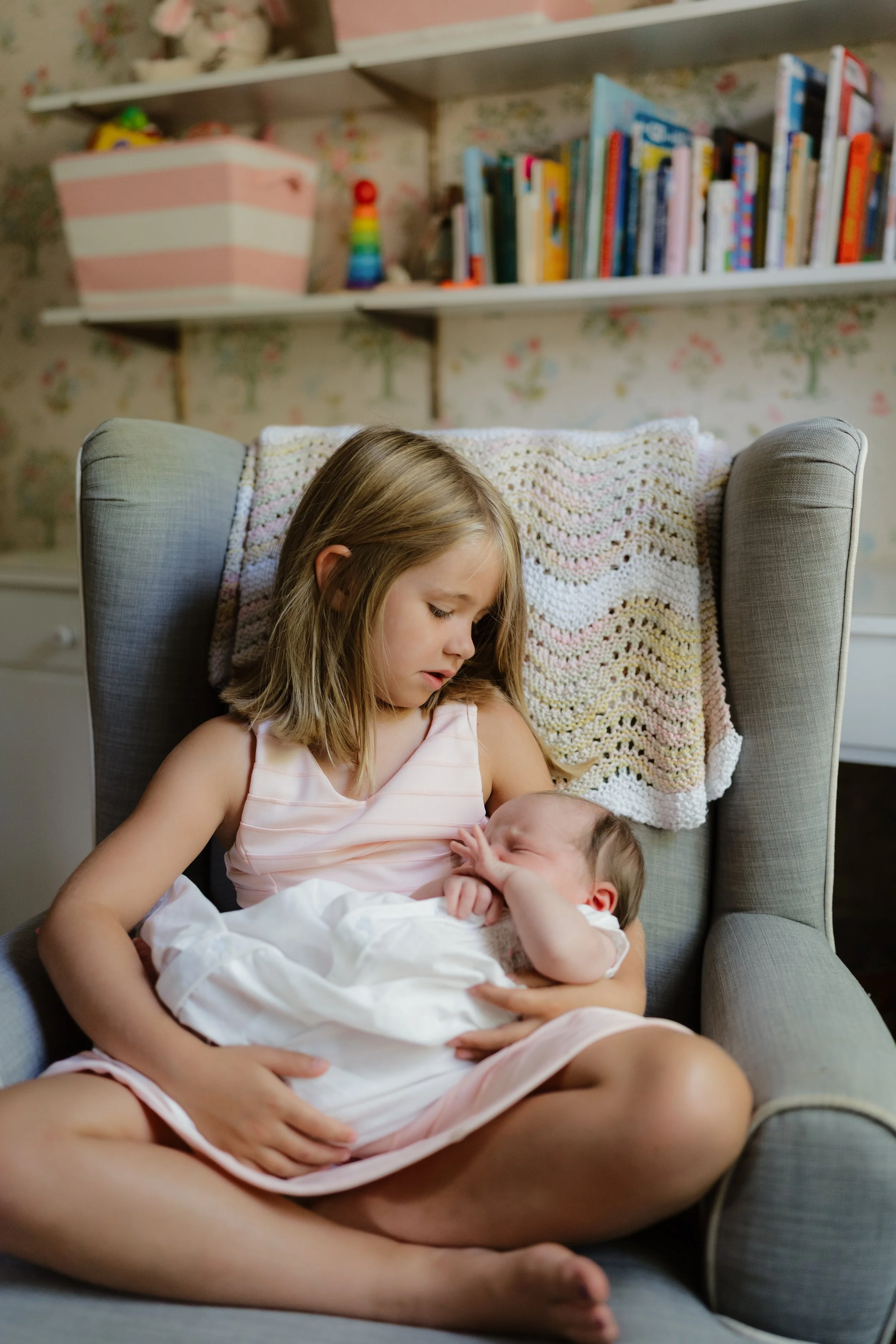 A young girl in a pink dress sitting in a gray armchair, holding and kissing a sleeping baby wrapped in white, with a crocheted blanket behind her in Memphis, TN. 