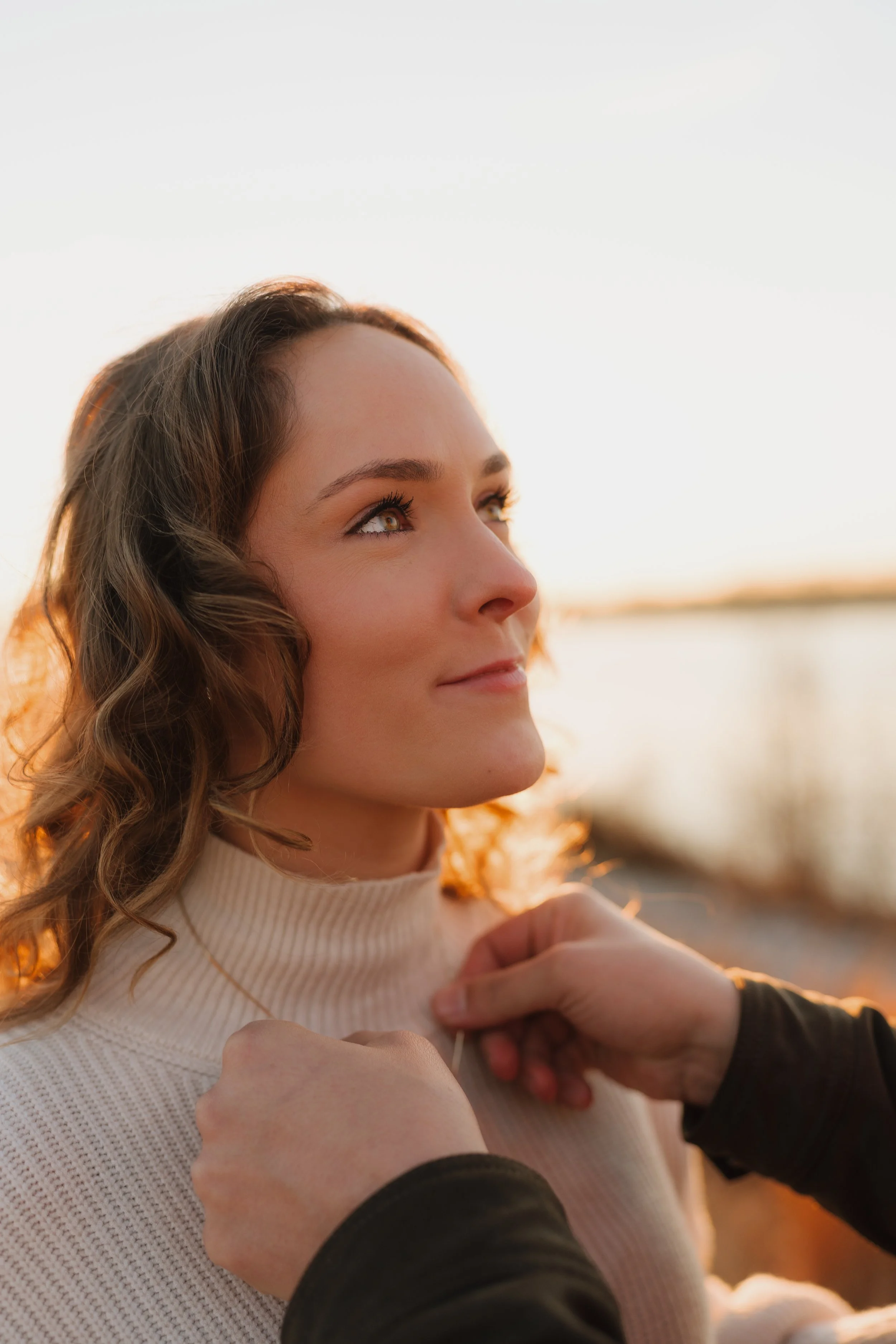 Close-up of an engaged woman with wavy brown hair and brown eyes, wearing a cream-colored ribbed turtleneck sweater, as someone assists her with a ring, with a blurred outdoor background during golden hour in Memphis, TN.