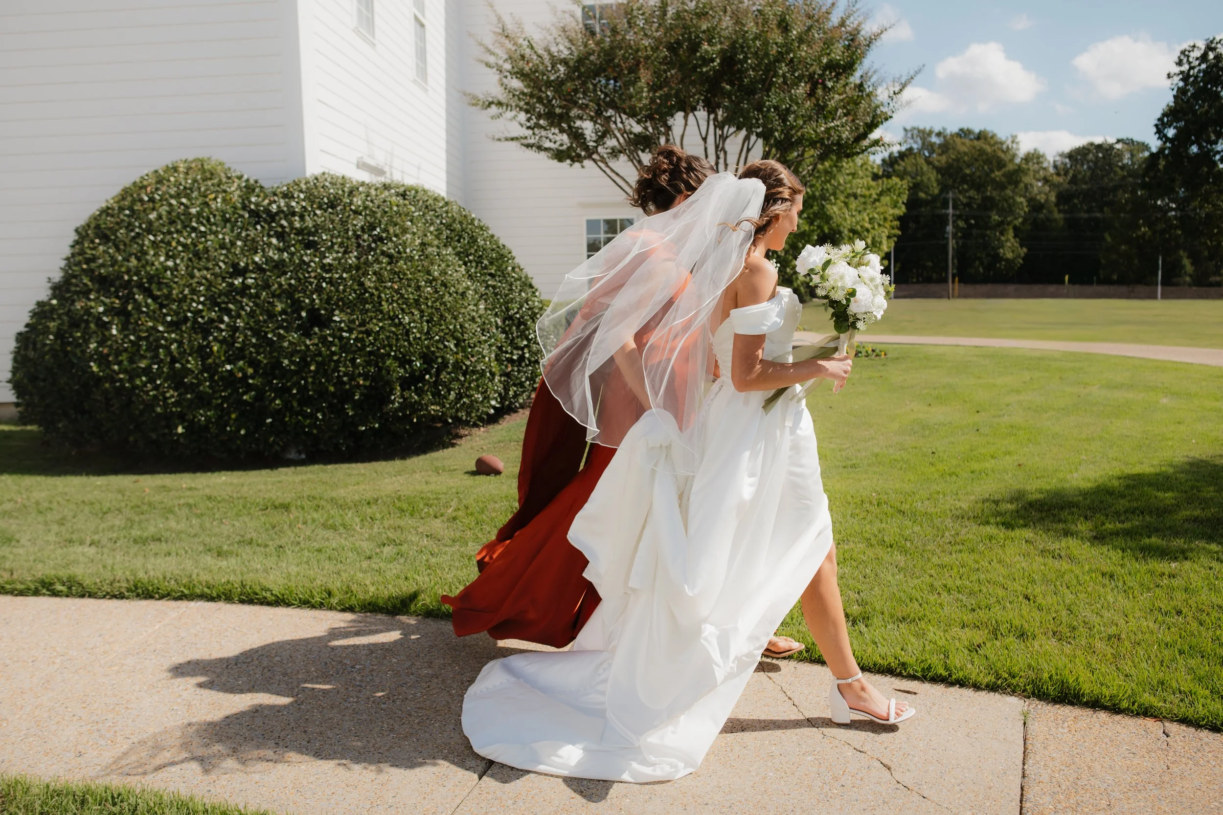 Bride in a white wedding dress and veil walking outside with a bridesmaid in a maroon dress and veil holding a bouquet of white flowers in Memphis, TN. 