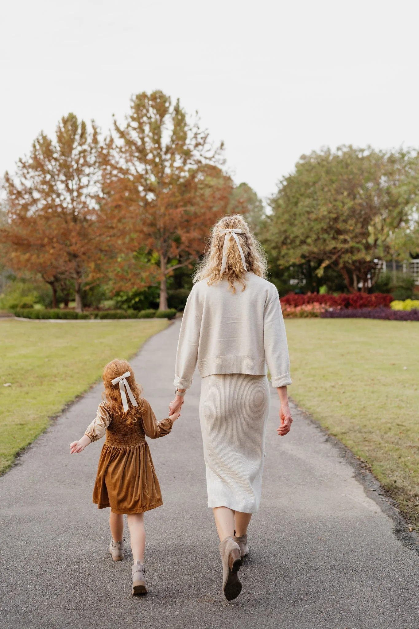 A mother and her daughter walk hand in hand down a paved path in a park with autumn trees and colorful bushes in Memphis, TN. 