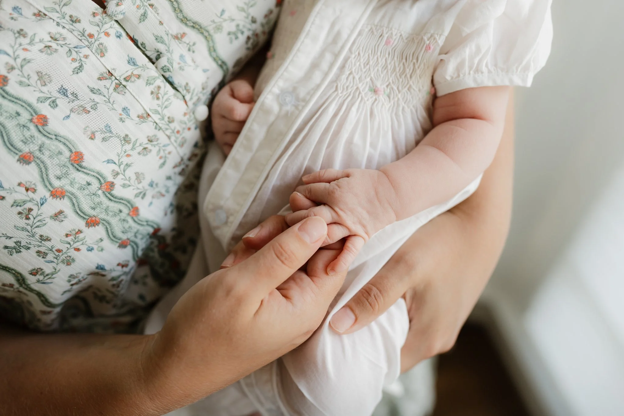 A close-up of a person holding a baby's hand, with the baby wearing a white dress with smocking details and embroidered pink and green flowers in Memphis, TN. 