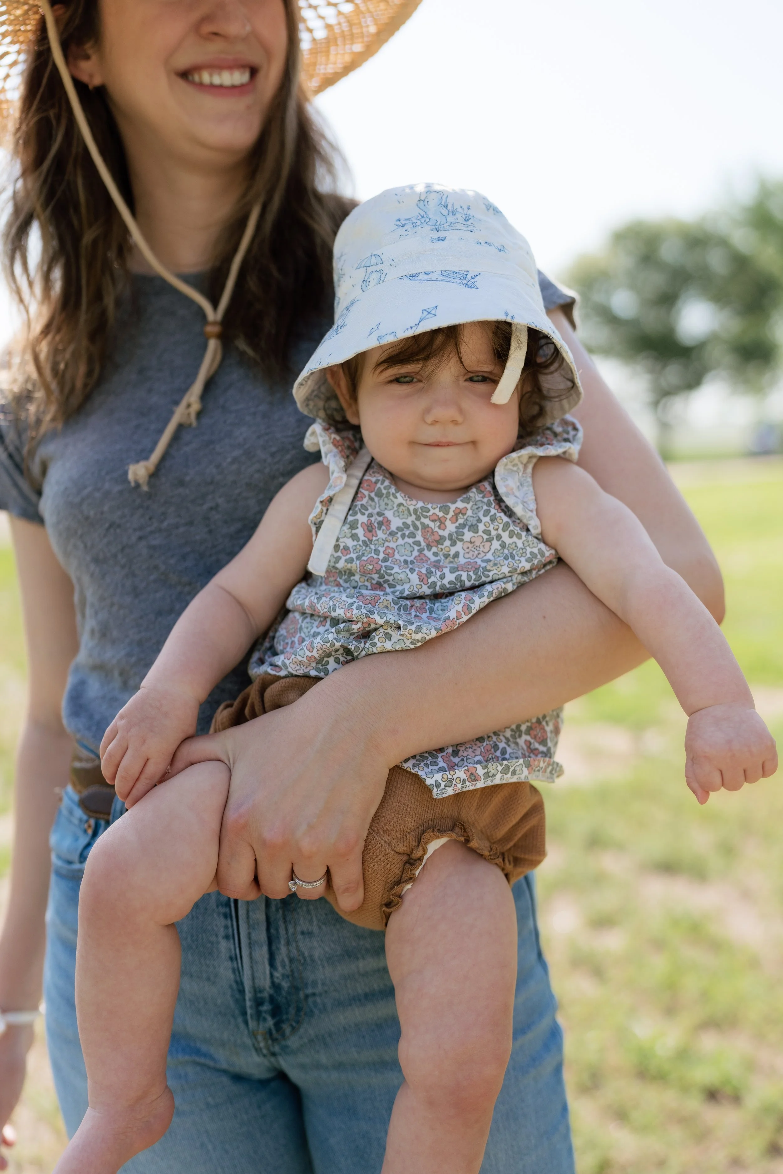A smiling woman in a large straw hat is holding up a young child in a floral-print sleeveless shirt and brown shorts outdoors on a sunny day, with green trees and grass in the background on a farm outside of Memphis, TN. 