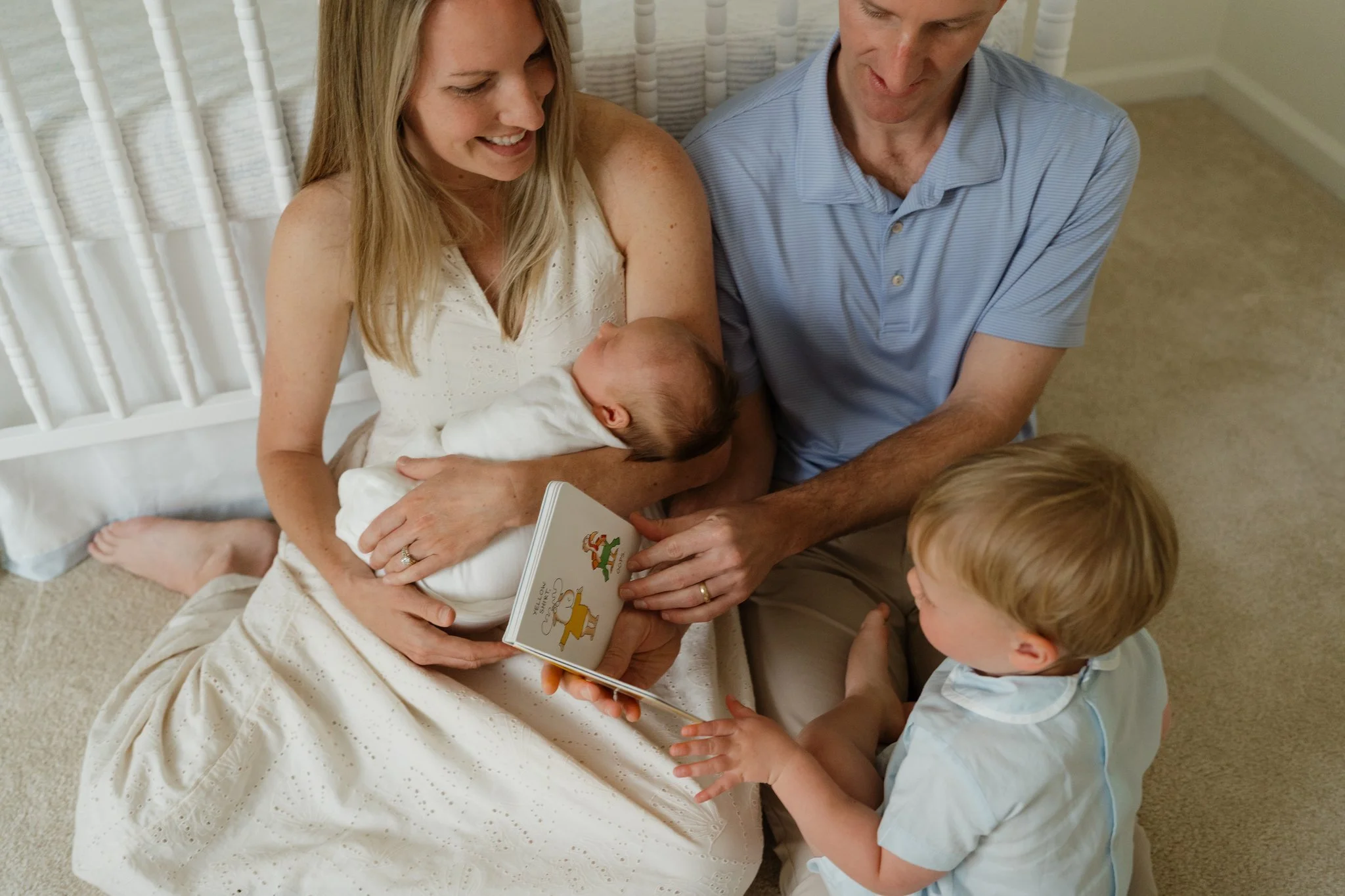 A woman with long blonde hair sitting on the floor holding a newborn baby, with a man and a young boy nearby. The woman and family members are looking at the baby and a book in Memphis, TN. 
