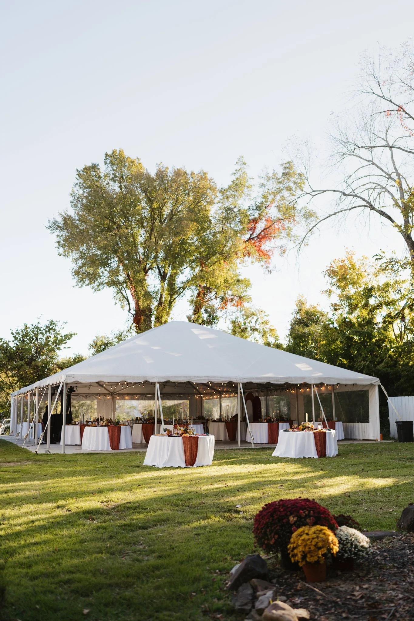 An outdoor event setup with a large white tent, decorated tables with white tablecloths and fall-colored accents, surrounded by fall flowers and trees with changing leaves, during daytime in Memphis, TN. 