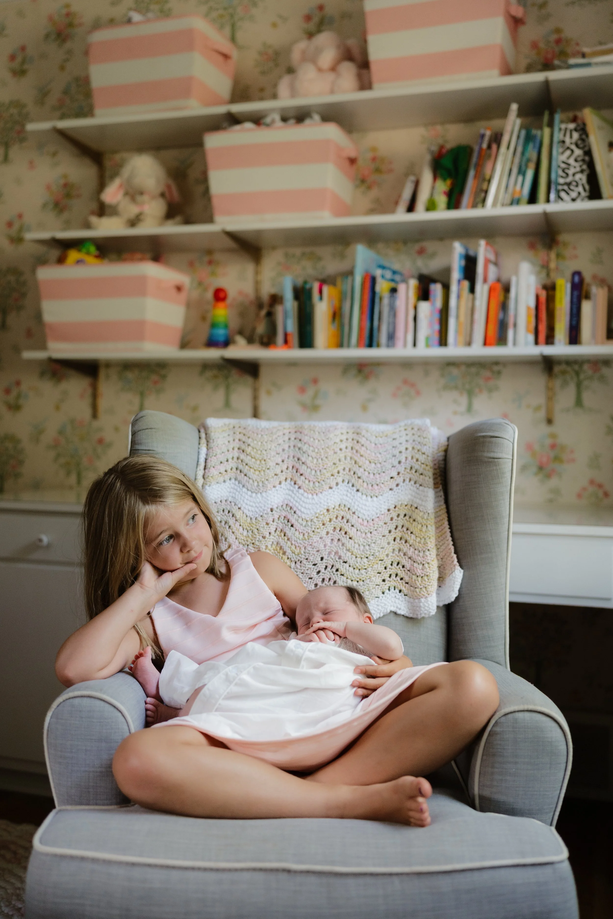 A young girl in a pink dress sitting in a gray armchair, holding a sleeping baby in her lap, in a room with a floral wallpapered wall and a white bookshelf with books and stuffed animals in the background  in Memphis, TN. 