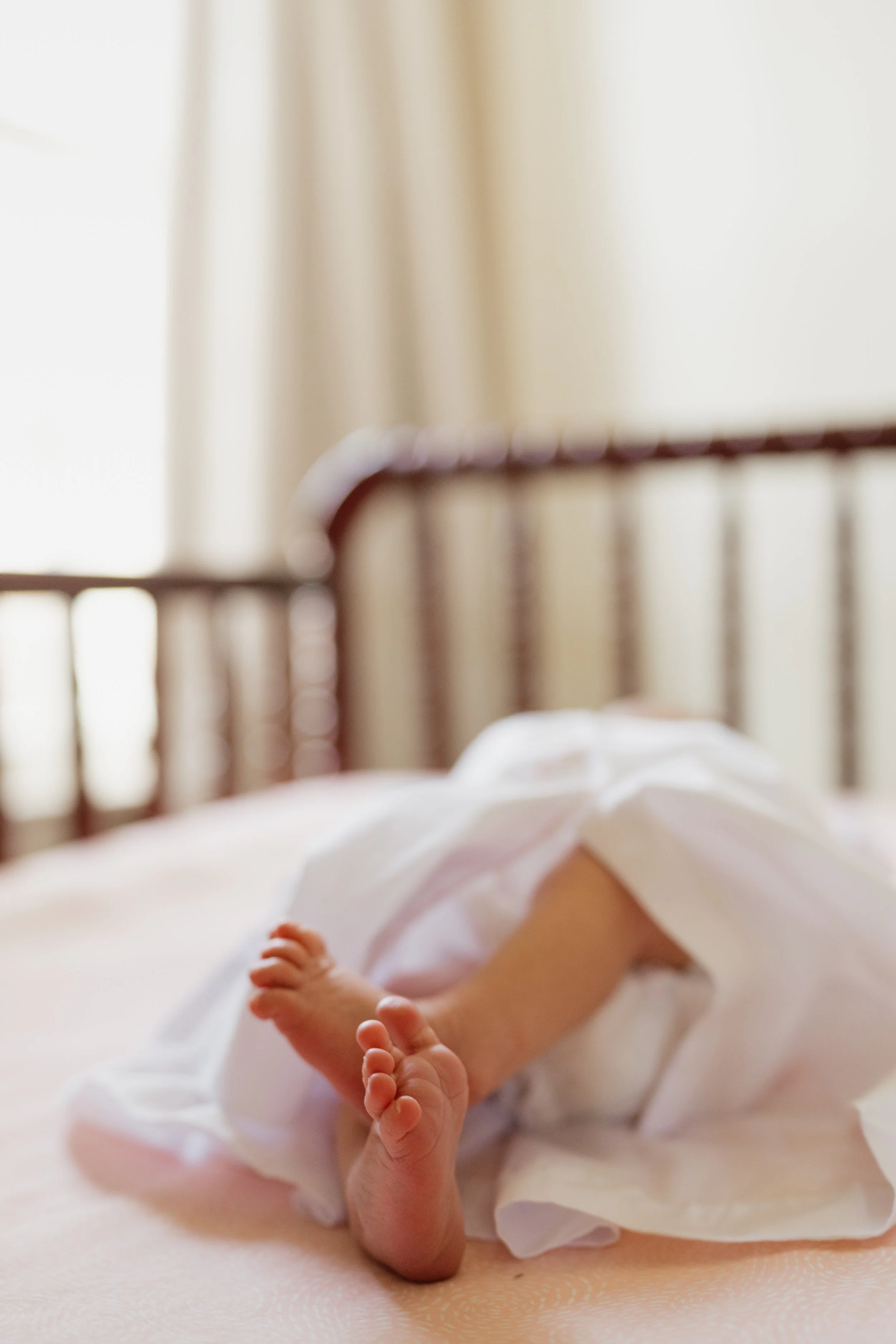 Close-up of a newborn baby's feet and hand resting on a soft surface, with a crib and light-colored curtains in the background in Memphis, TN. 