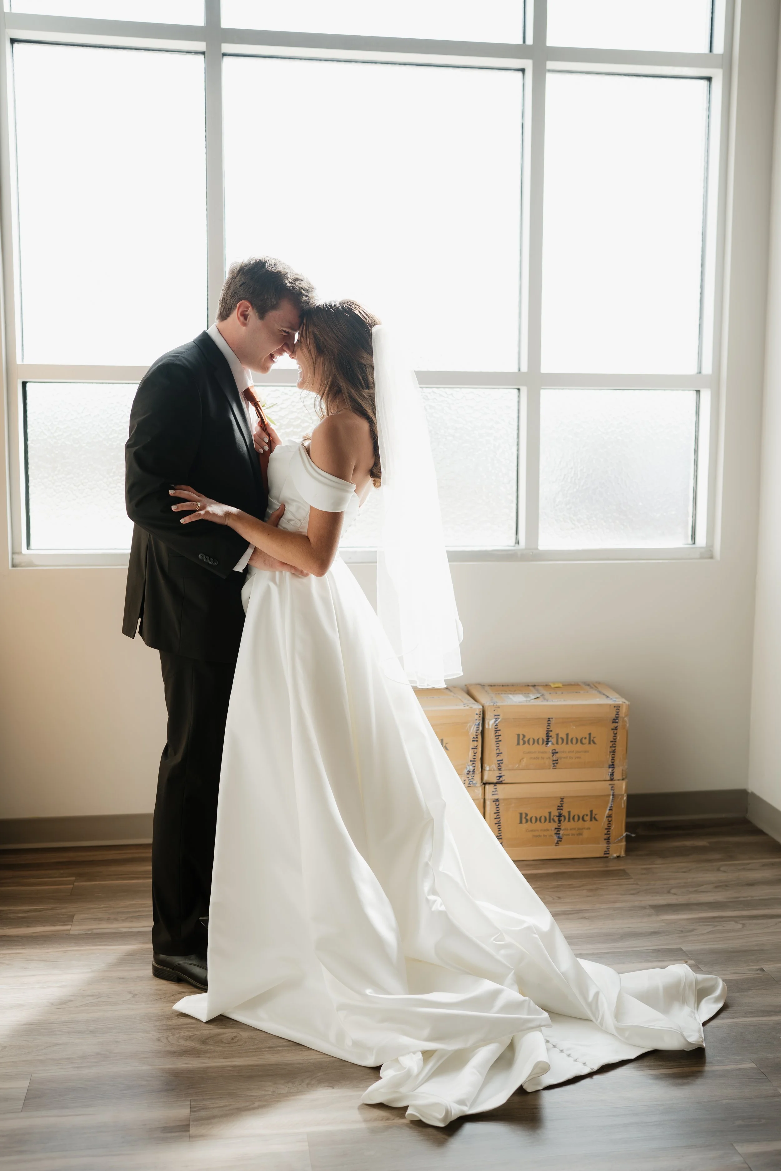 A bride and groom are touching foreheads and smiling in front of a large window, with wedding attire and moving boxes nearby in Memphis, TN. 