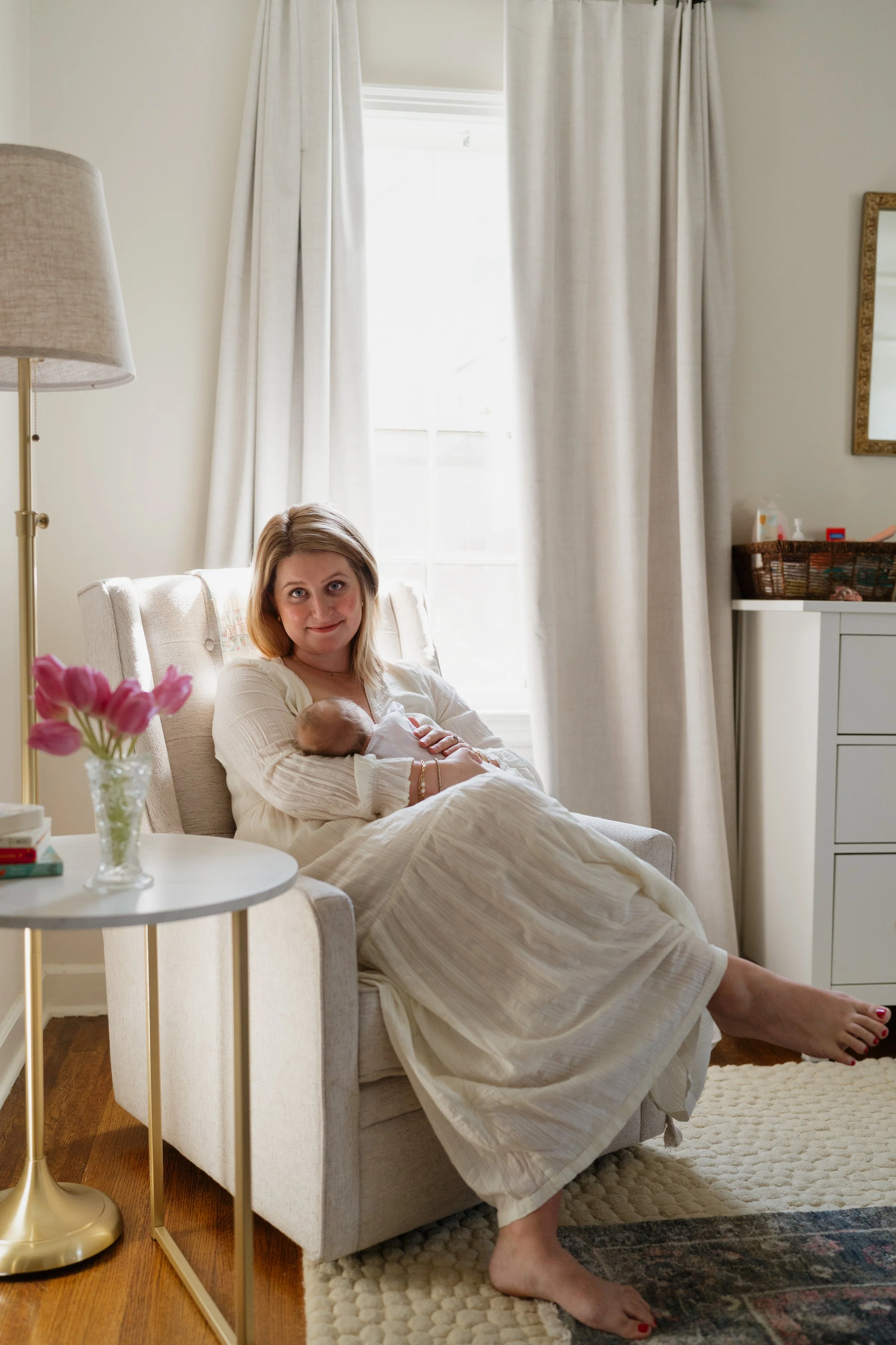 A woman with blonde hair sitting on a cream-colored armchair, cradling a newborn baby. She is wearing a white dress and looking at the camera with a gentle expression. The room has cream curtains, a small side table with pink flowers, and a white dre
