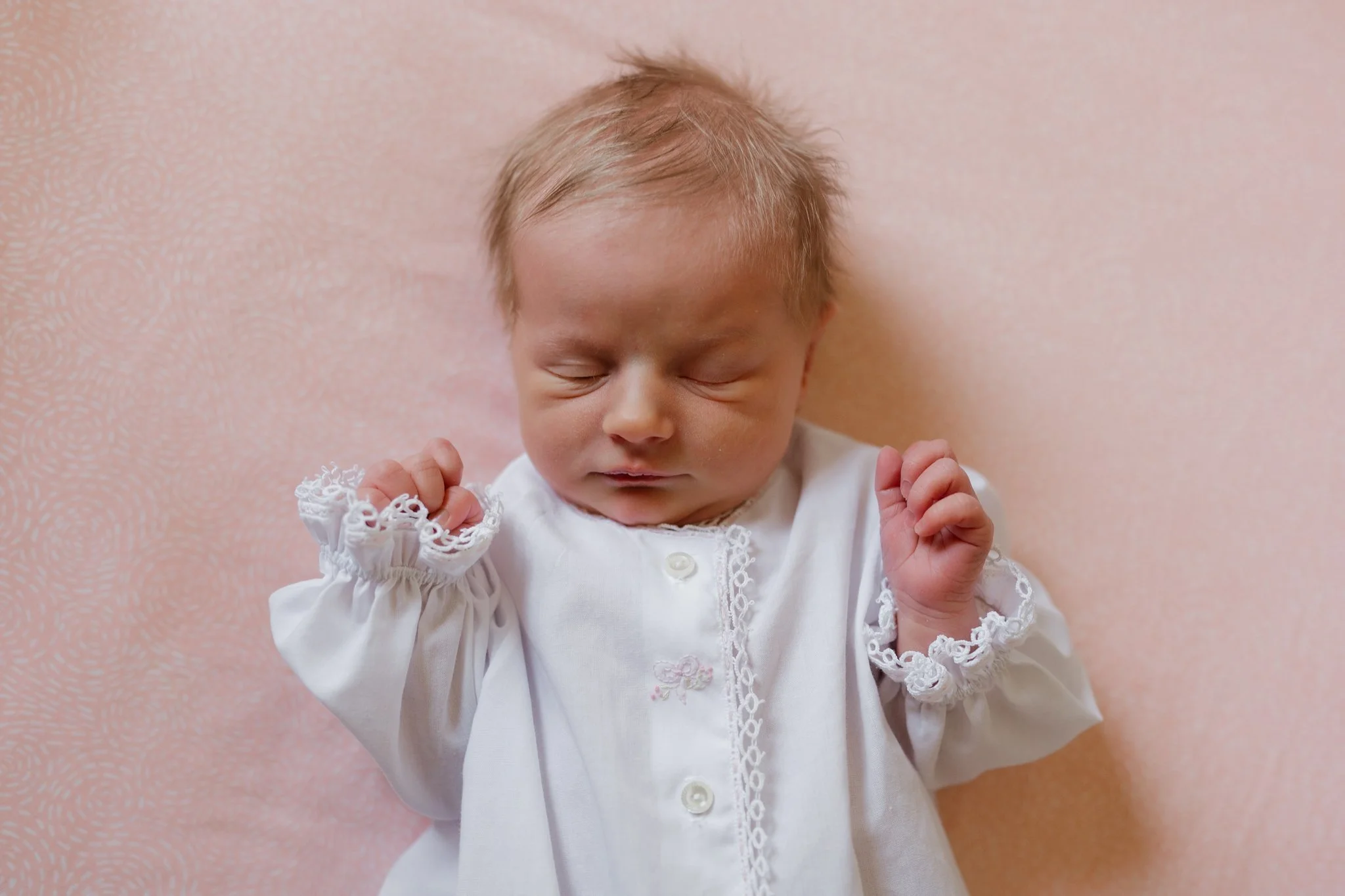 A baby with closed eyes, wearing a white dress with lace details, has their fists clenched and raised slightly, against a pink textured background.
