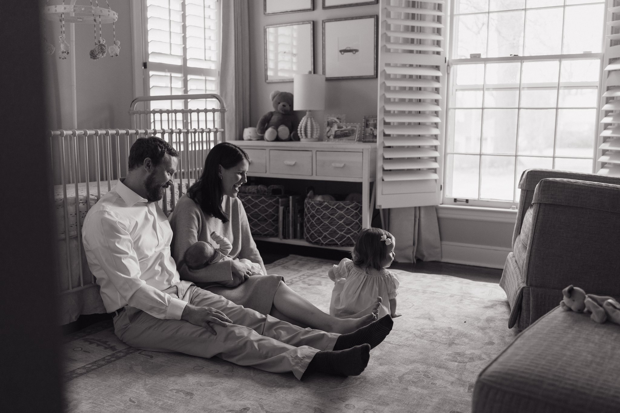 A family sitting on the floor in a sunlit living room, with a man, woman, and a toddler girl engaging with a plush teddy bear and toys. The room has large windows with shutters, framed pictures on the wall, and various toys and decor in Memphis, TN. 