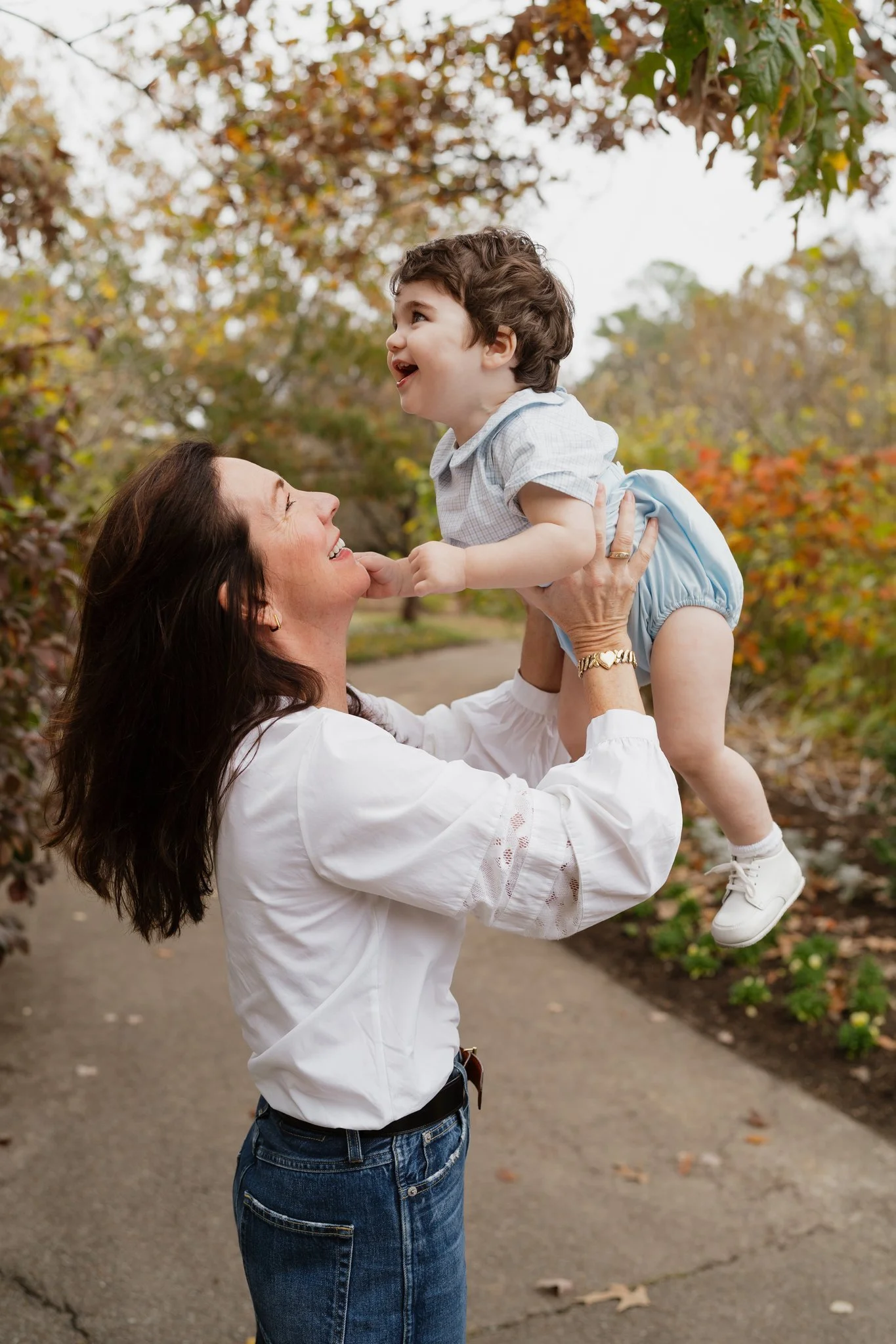 A woman is lifting a young boy, who appears to be happy and laughing, outside on a fall day with trees and fallen leaves in the background in Memphis, TN. 