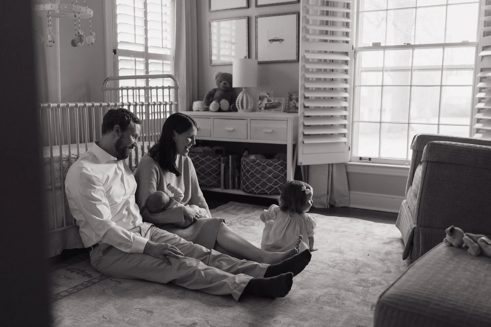 A black-and-white photo showing a man, woman, and young girl sitting on a rug inside a well-lit nursery. The man and woman are sitting close together, holding hands, and looking at a toy dog. The girl is sitting in front of them, facing a stuffed ani