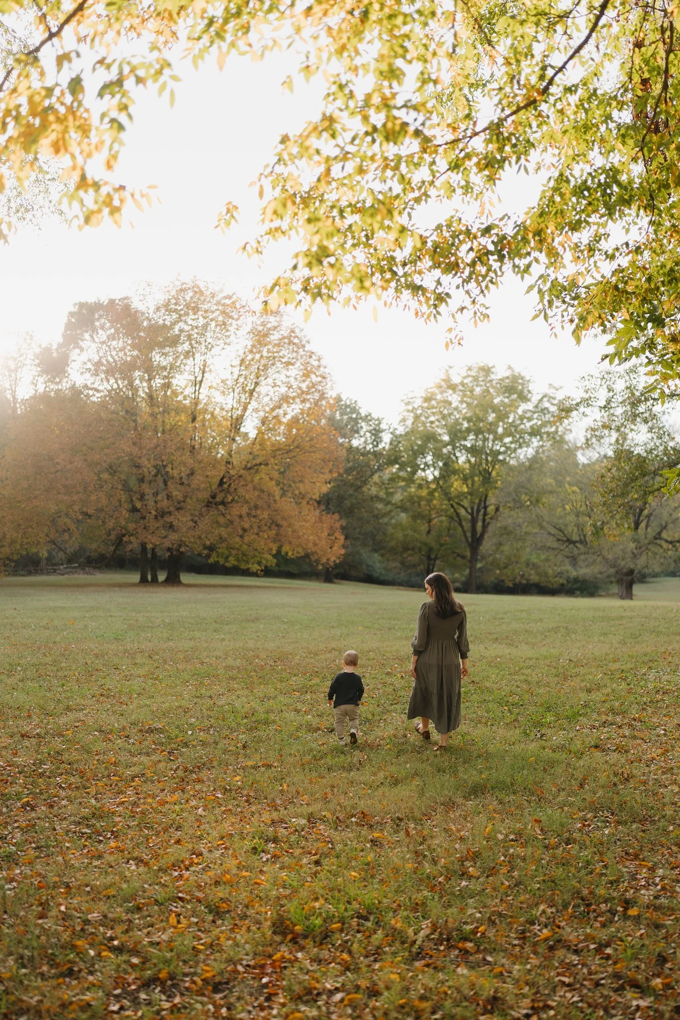 A woman and a young boy walking away from the camera in a park during autumn. The ground is covered with fallen leaves, and the trees have autumn-colored foliage. The sky is bright and slightly overexposed in Memphis, TN. 