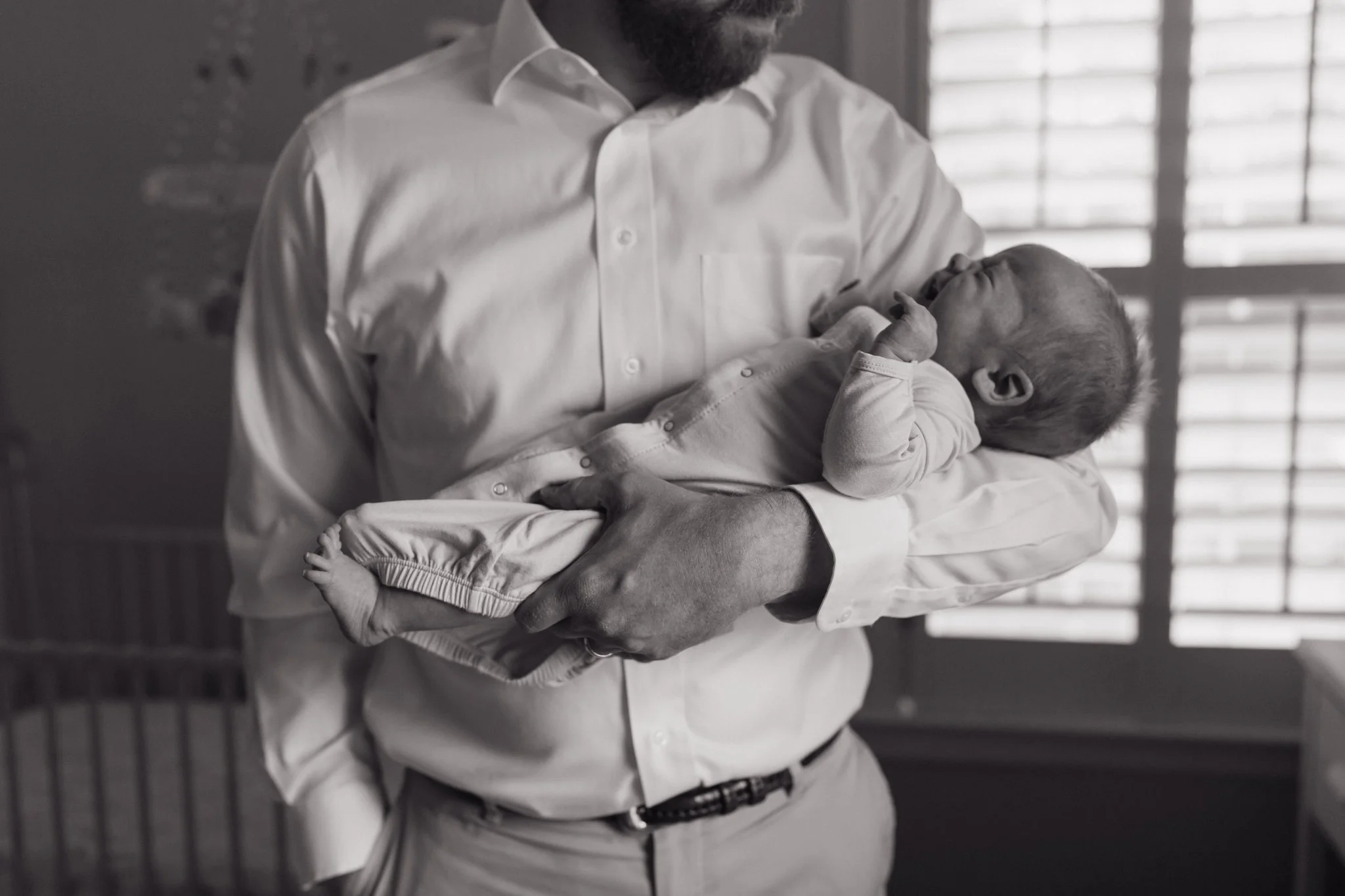 A man wearing a white button-up shirt holding a baby in his arms indoors, with a window with blinds in the background in Memphis, TN. 