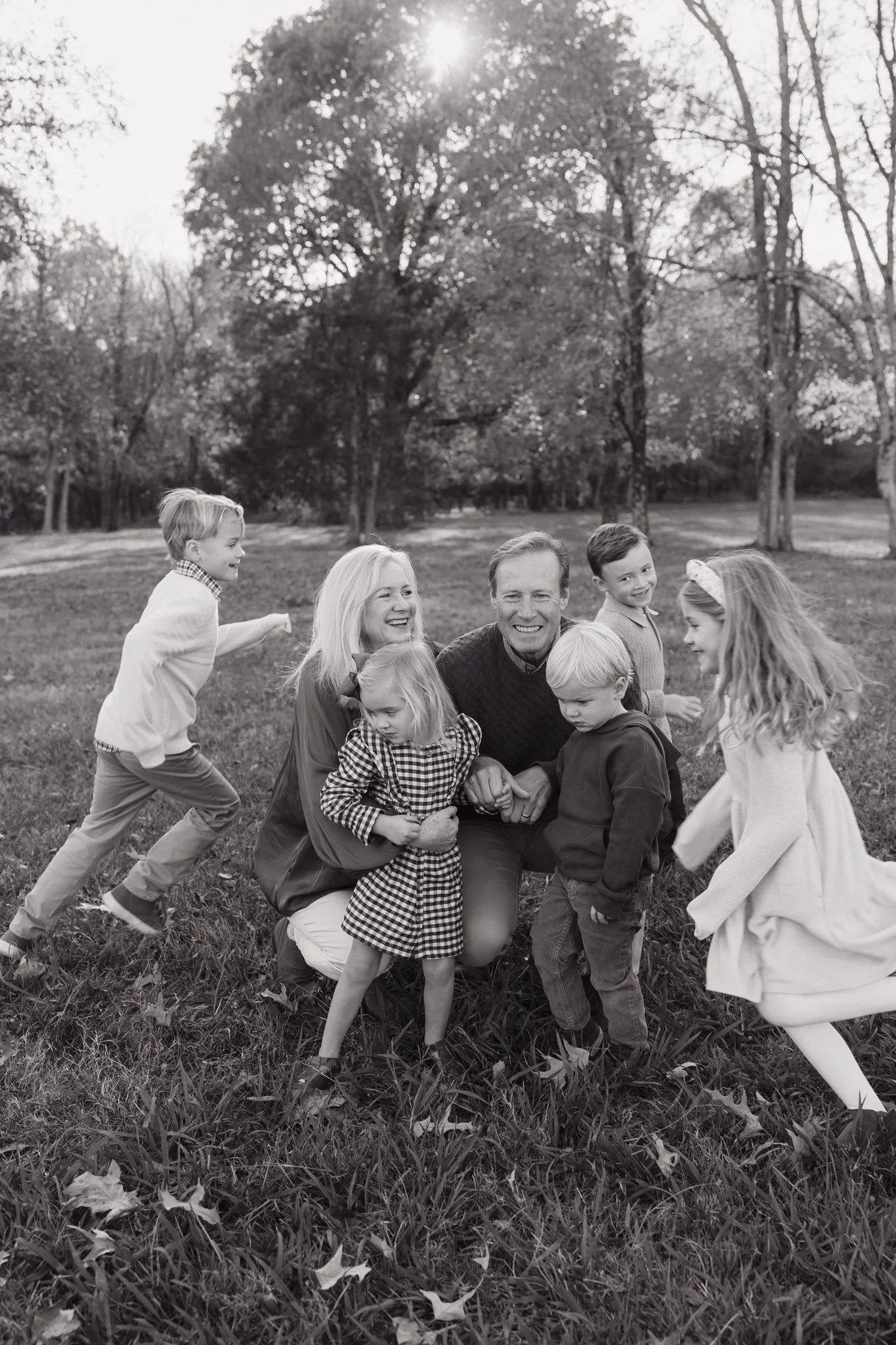A black and white photo of a smiling family of adults and children outdoors in a park. The family is playing and interacting on the grass with trees and sunlight in the background in Memphis, TN. 
