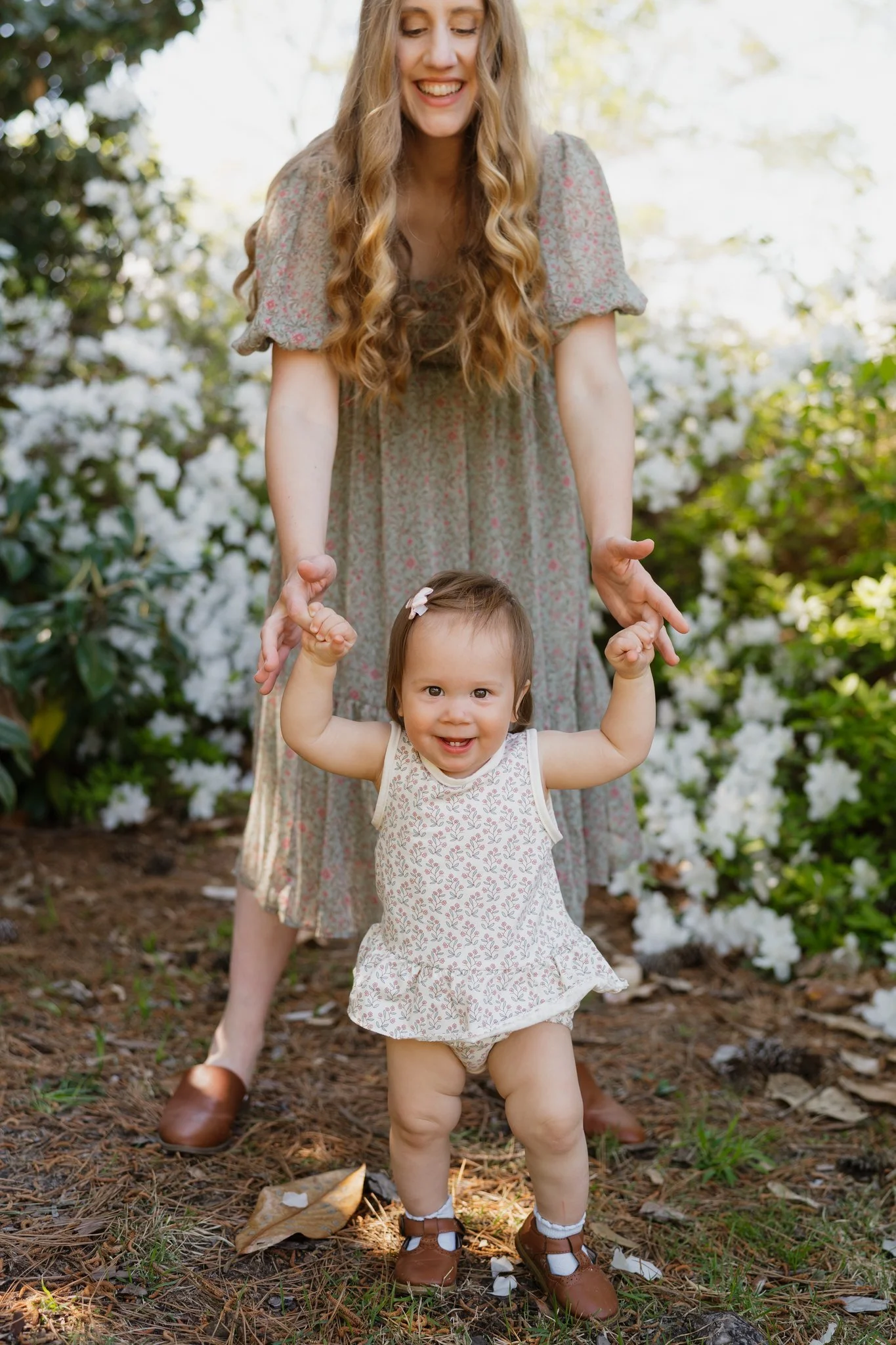 A woman with long blonde hair smiling and standing behind a young girl with short brown hair, who is excitedly holding hands raised in the air. They are outdoors among blooming white flowers and green foliage on a sunny day in Memphis, TN. 