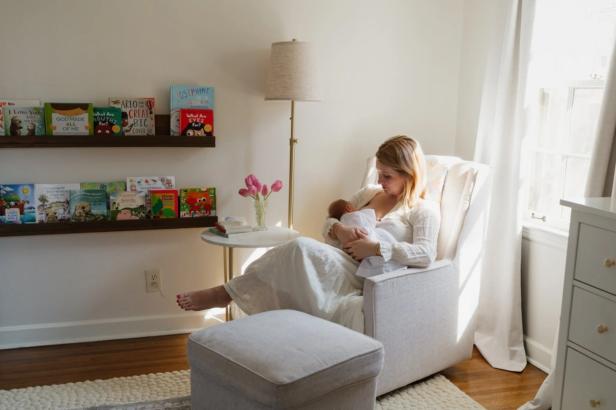 A woman sitting on a white armchair breastfeeding a baby in a bright room with sunlight streaming through a window. There are books and a vase with pink flowers on a small round table beside her, and a wooden shelf with children's books on the wall i