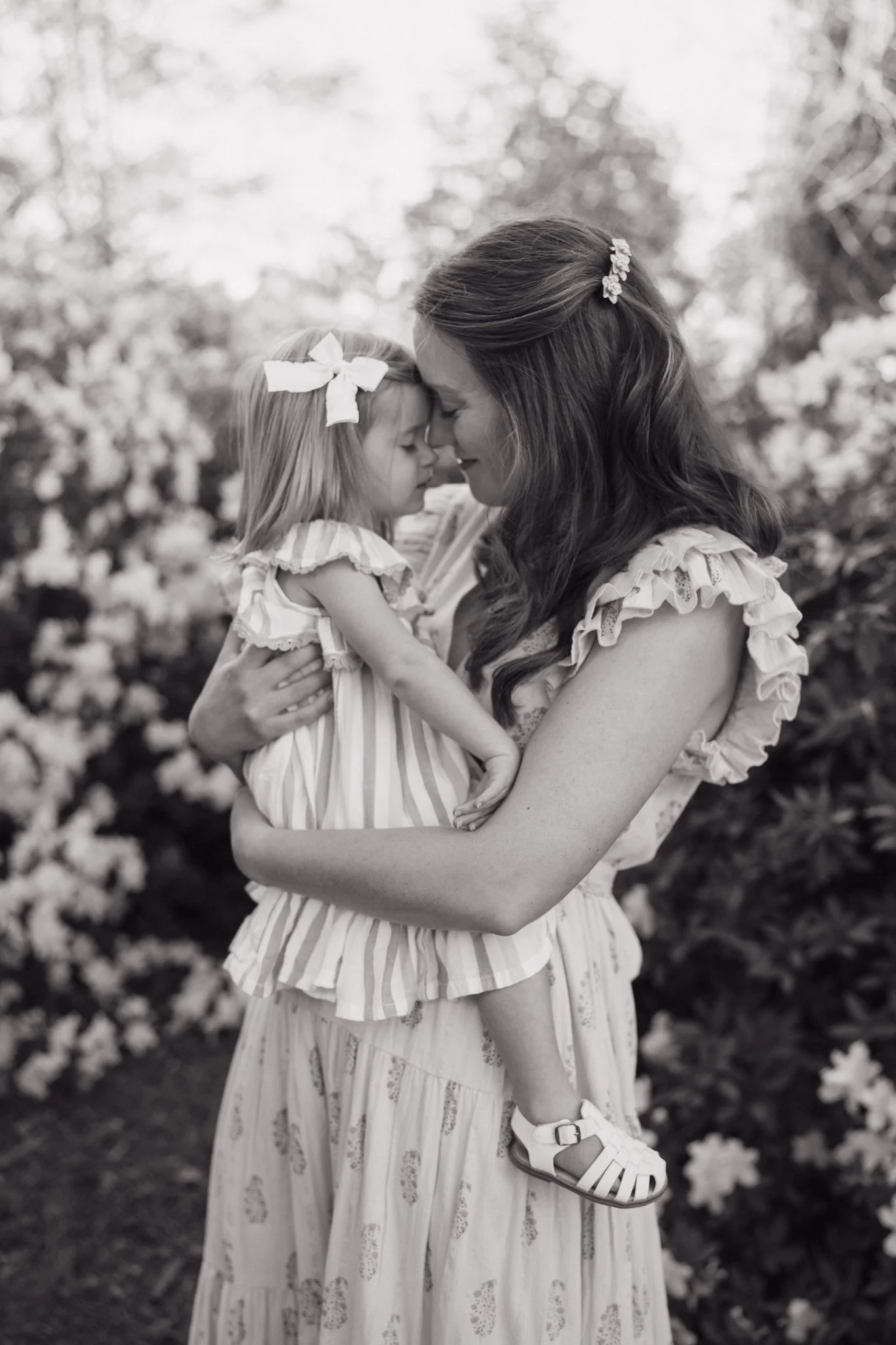 A woman and a young girl are holding each other with their foreheads touching in a close, tender pose outdoors, with blurred flowers in the background. They are wearing light-colored, ruffled dresses and appear to be sharing an intimate moment in Mem