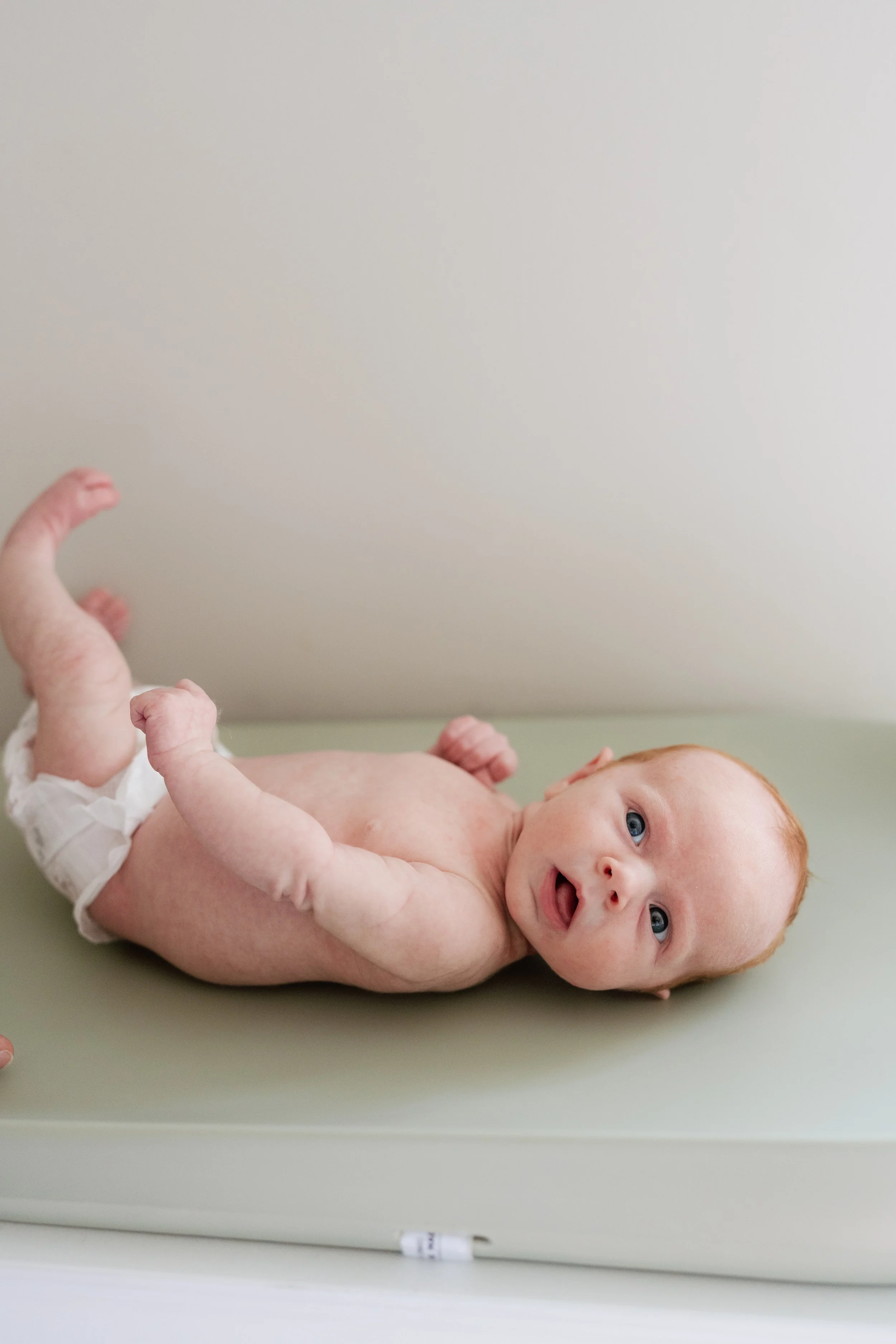 A baby lying on their back on a green changing table with a plain light-colored wall behind them, looking at the camera with blue eyes and open mouth, with some hair and skin showing, and wearing a white diaper in Memphis, TN. 