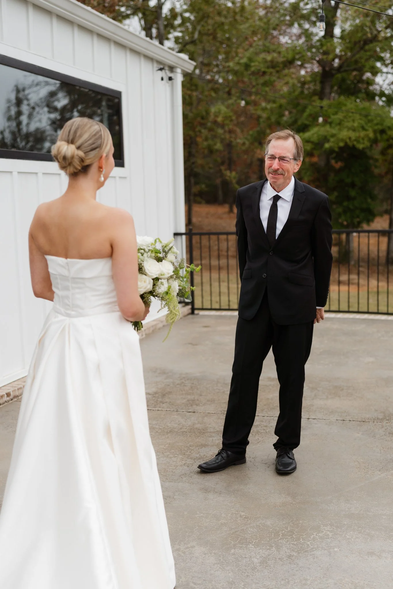 A bride in a strapless white wedding dress holding a bouquet of white flowers stands outdoors, smiling at an older man wearing glasses and a black suit, who is also smiling. They are near a white building with a black fence and trees with green and o