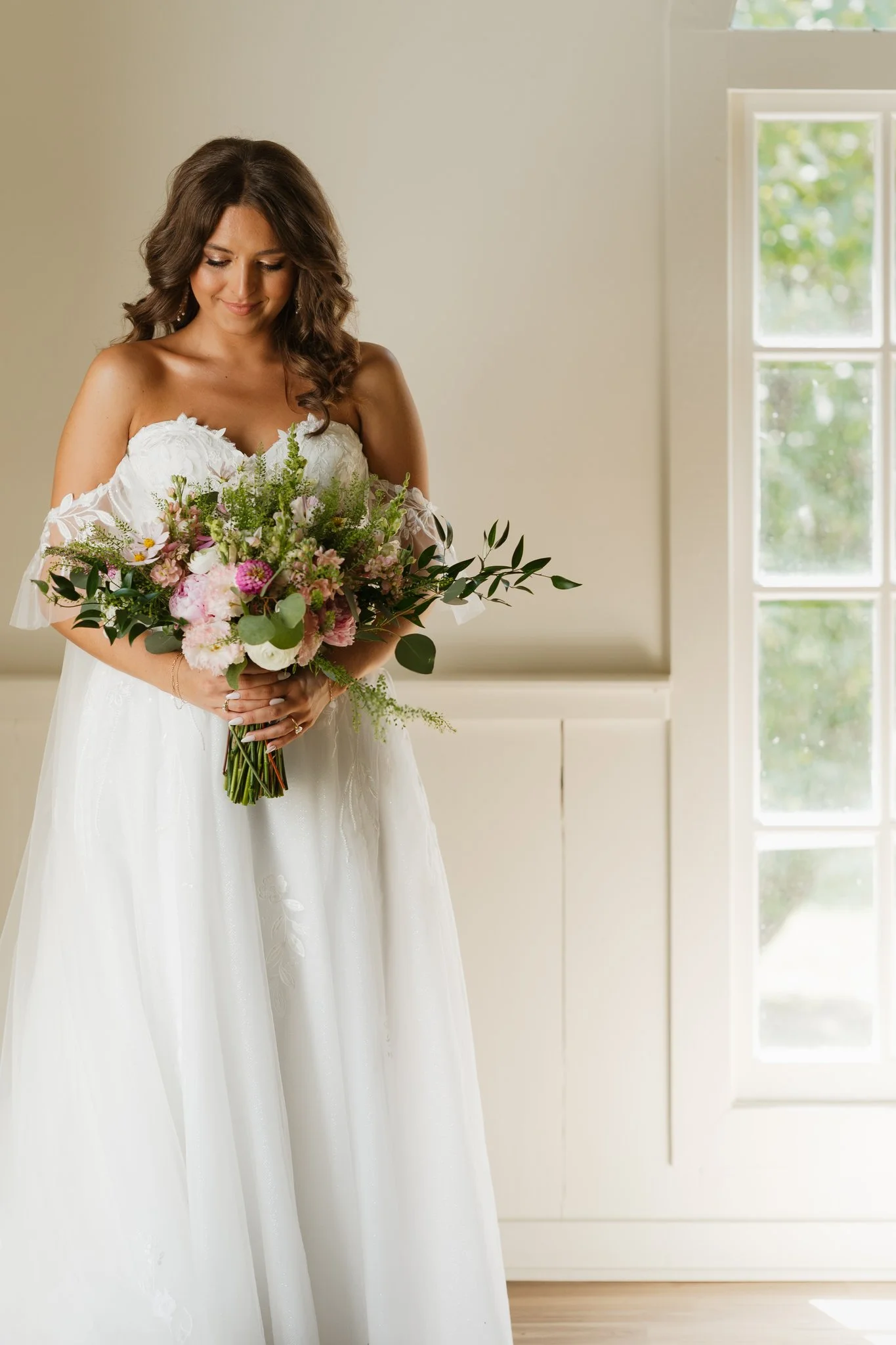 A woman in a white wedding dress holding a bouquet of pink and white flowers, standing near a window with greenery outside in Memphis, TN. 