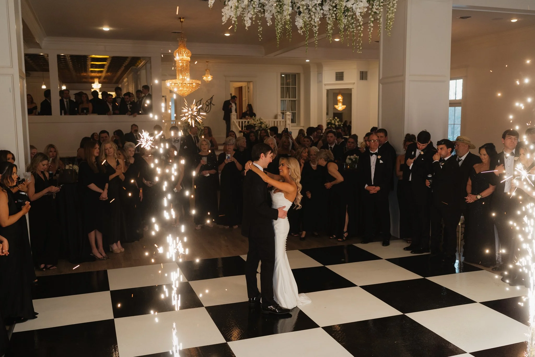 A wedding reception with the bride and groom dancing in front of a crowd, surrounded by sparklers and string lights, elegant chandeliers, and floral decorations in a bright venue in Memphis, TN. 