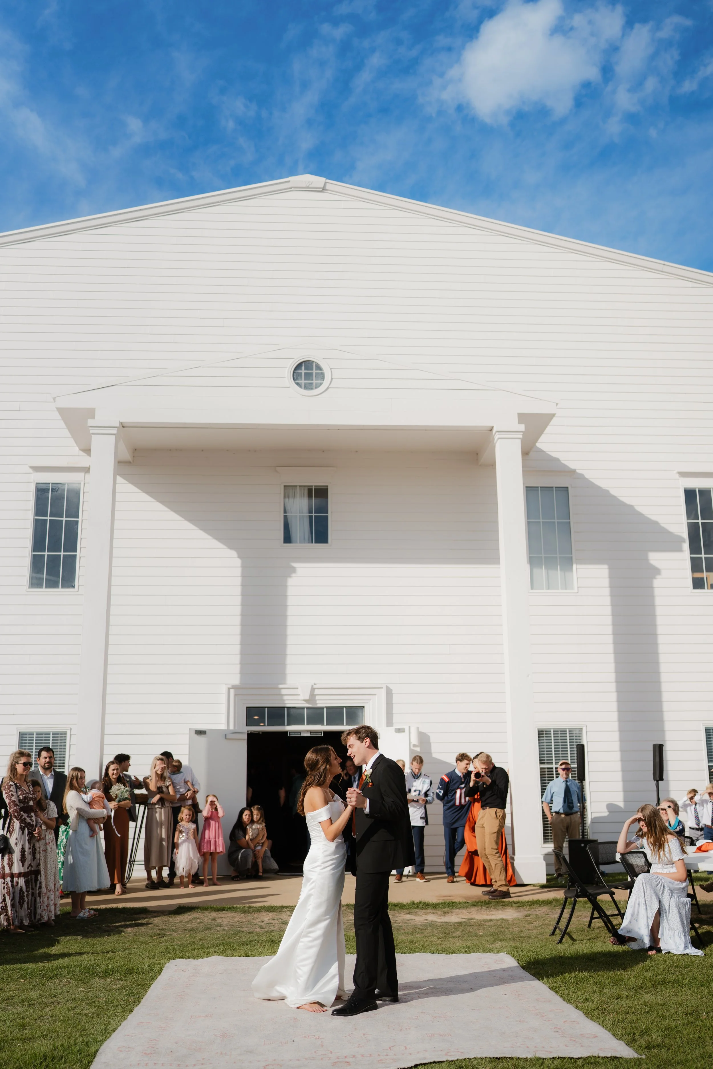 A wedding ceremony outdoors in front of a white church with a gathering of guests. The bride and groom are dancing on a small carpeted area, holding hands and looking at each other. The bride is in a white wedding dress, and the groom is in a black s