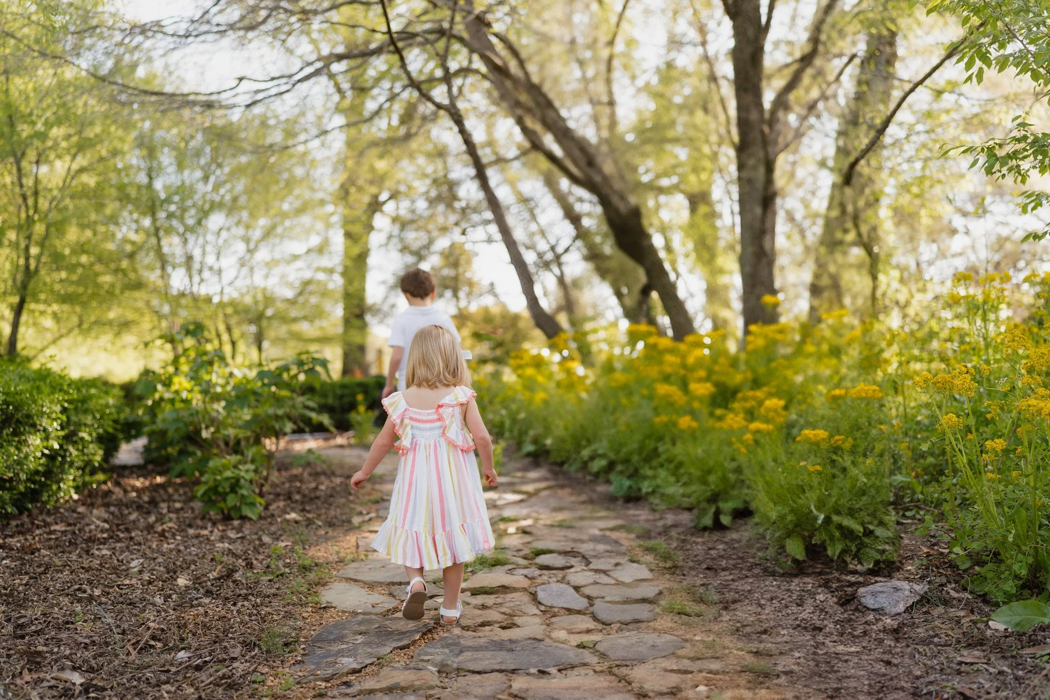 Two children, a girl and a boy, walking on a stone path through a lush green garden with trees and yellow flowers in Memphis, TN in Memphis, TN. 