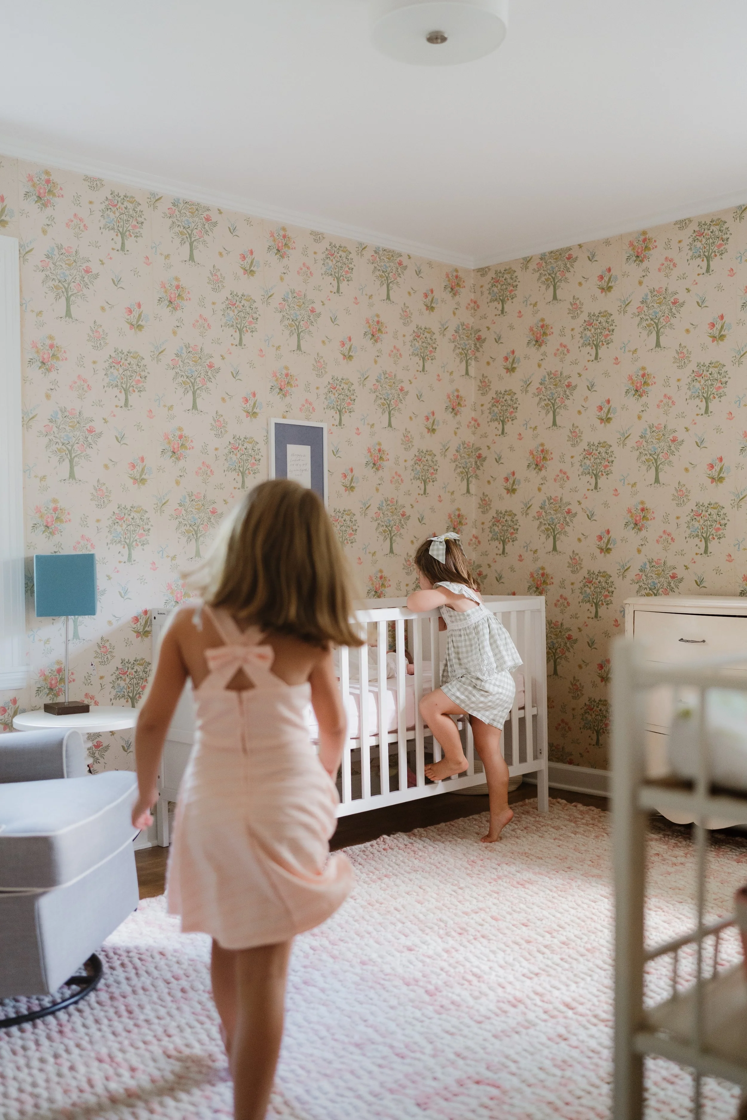 Two young girls are in a bedroom with floral wallpaper. One girl with shoulder-length blond hair in a pink nightgown is walking, and a second girl with long brown hair tied with a bow is climbing into a white crib. The room has a pink carpet, a blue 