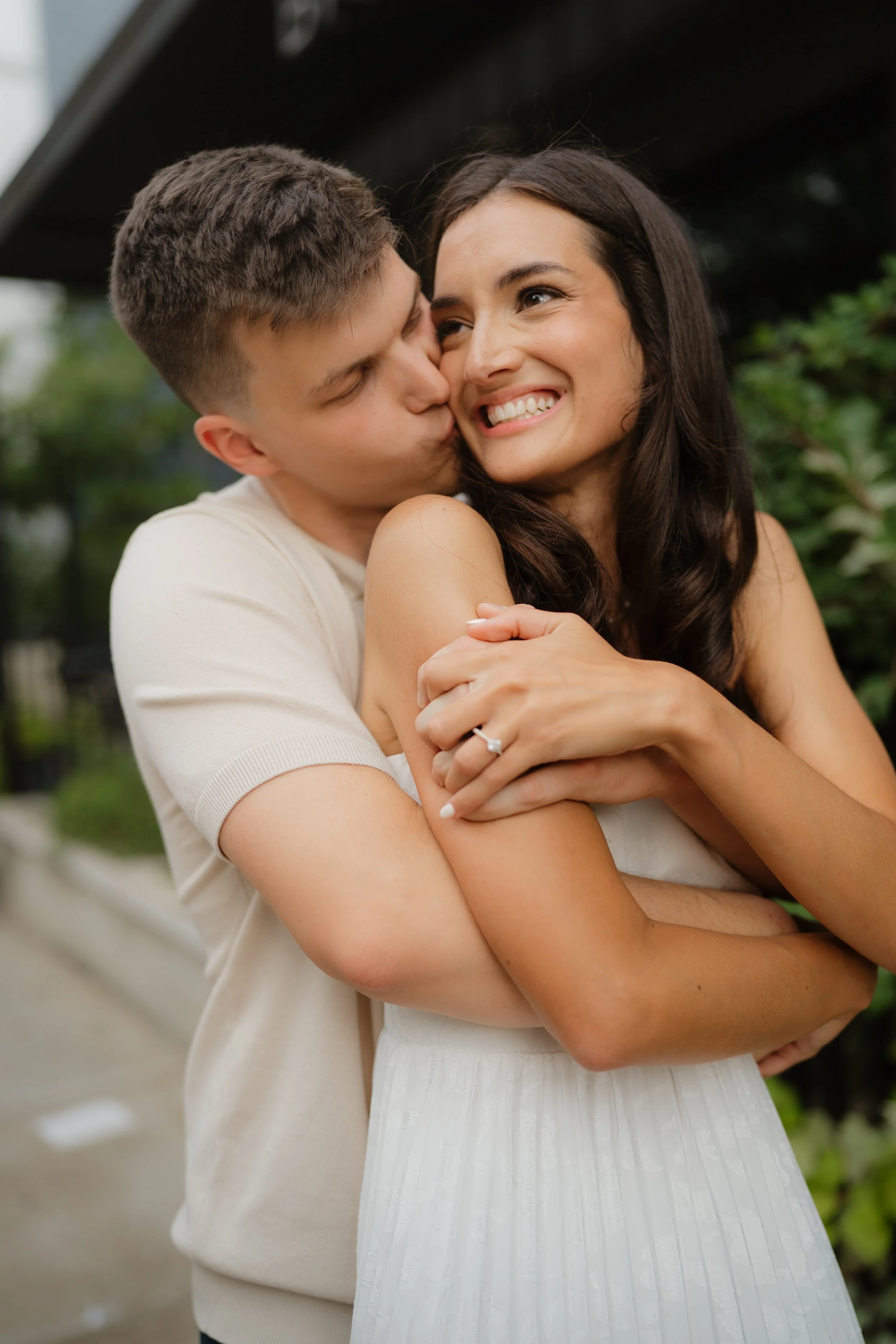 A young man kissing a woman on the cheek outdoors while hugging her. The woman is smiling and wearing a white dress. The man is wearing a beige shirt. A beautiful engagement shoot in Memphis, TN. 