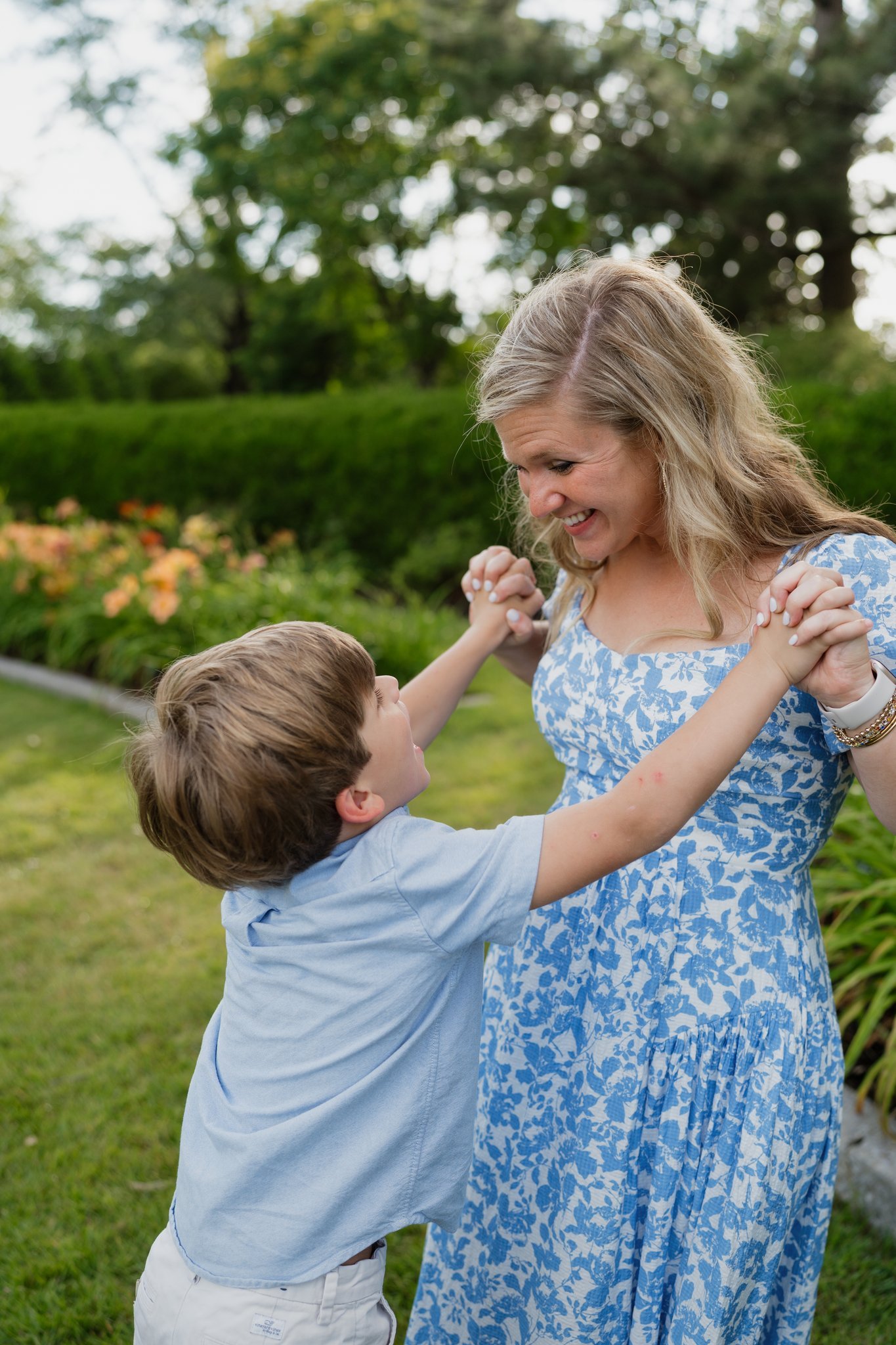 A woman and a young boy dancing outdoors in a garden with trees and flowers in the background.
