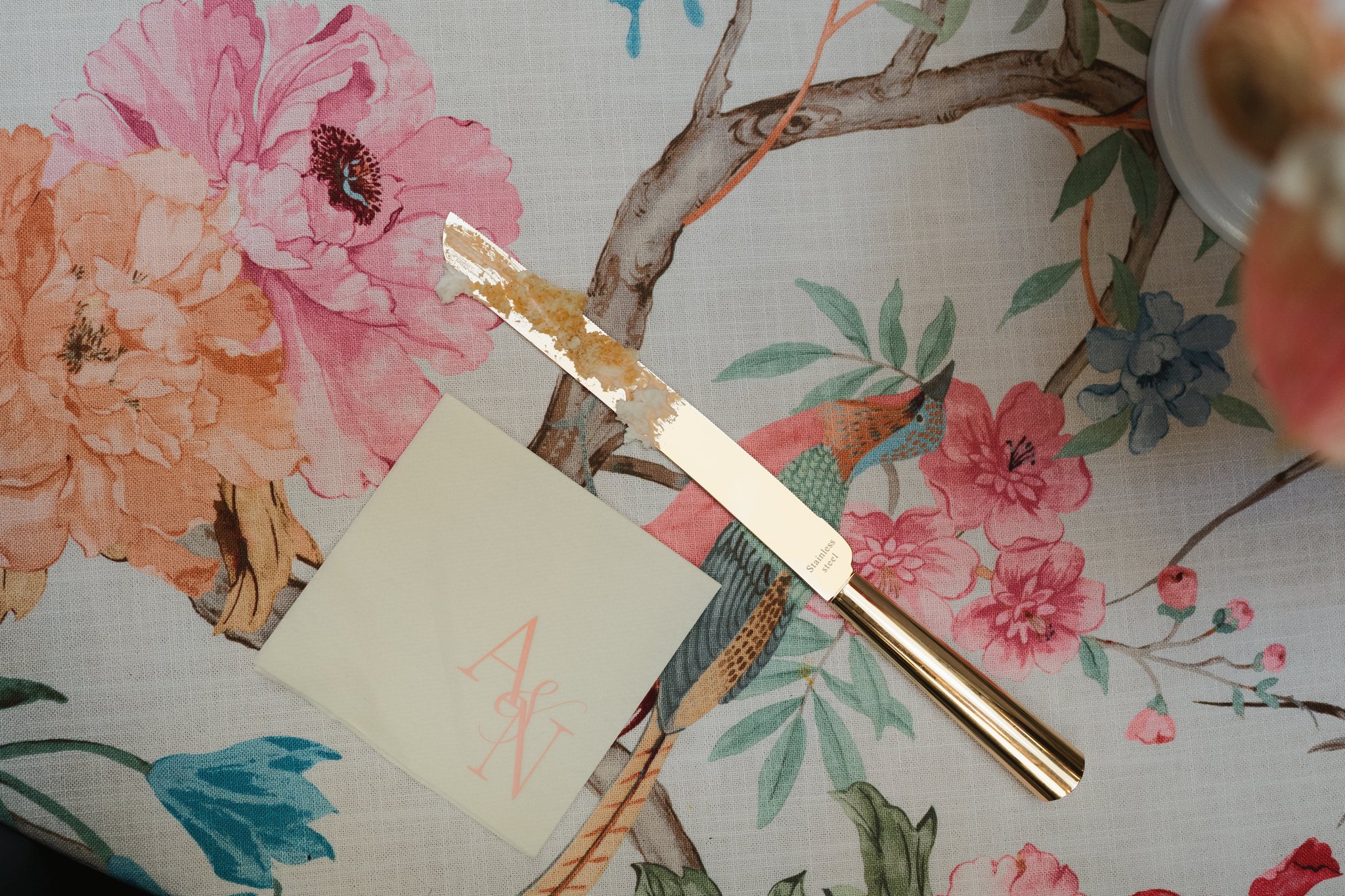 A gold-colored stainless steel pen with a patterned black and white grip, placed on a floral tablecloth with peony and other flowers, beside a white sticky note, a beige card with pink writing, and part of a blurred object on the top right in Memphis