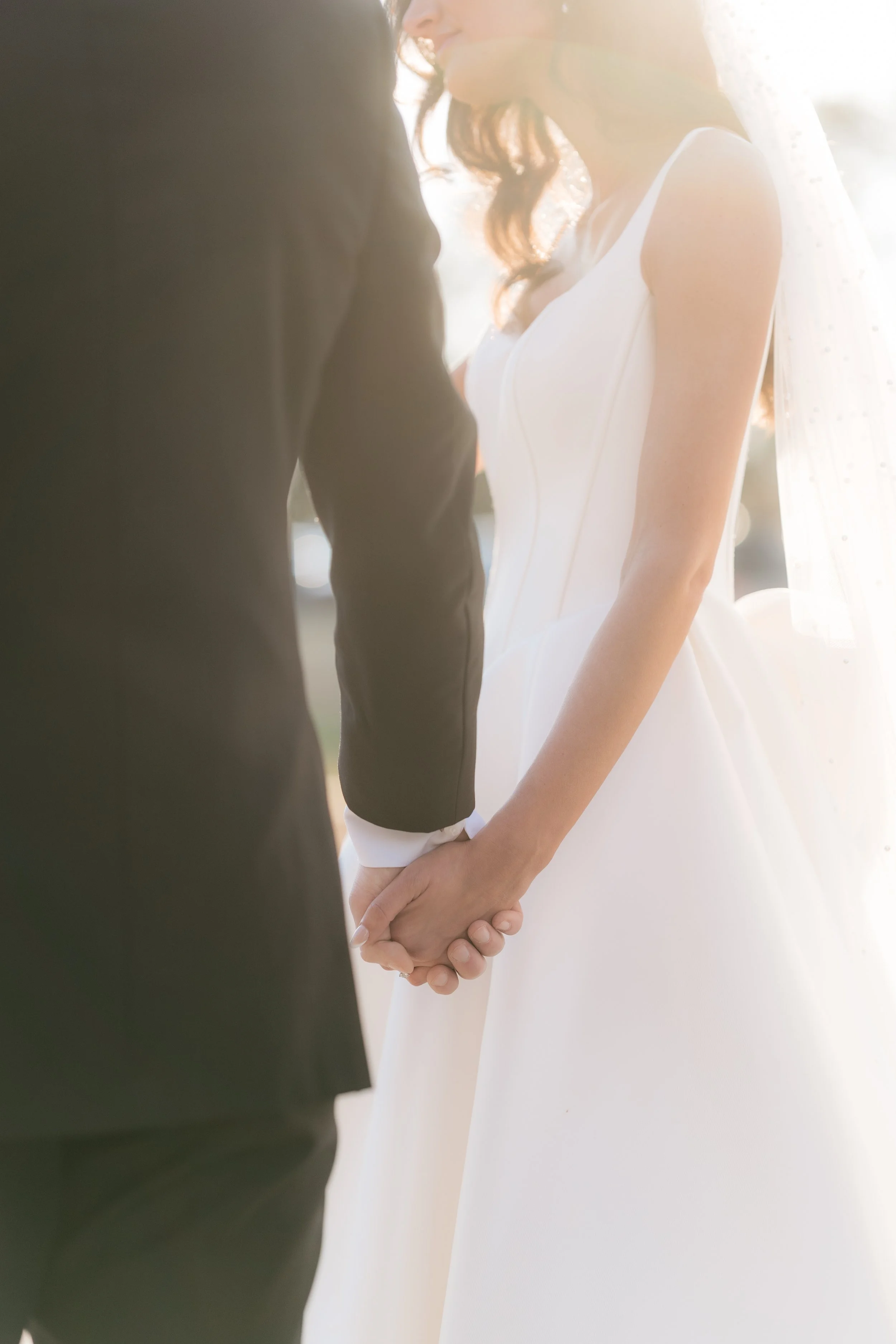 A bride and groom are holding hands during their wedding ceremony, with the bride wearing a white wedding dress and veil, and the groom in a dark suit. The image captures a close-up of their hands and part of their upper bodies, with sunlight creatin