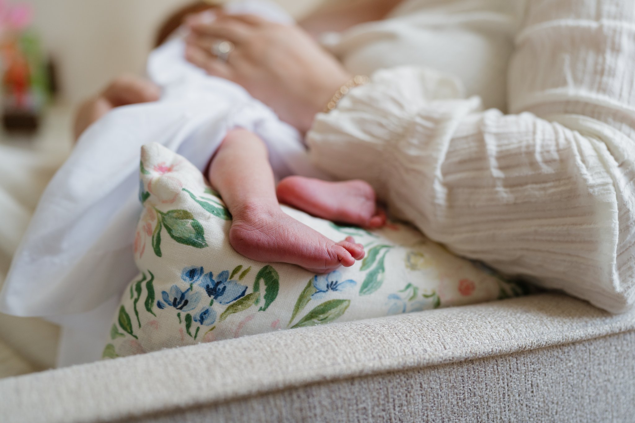Close-up of a baby's hand holding an adult's finger, lying on a floral blanket. The scene suggests a tender moment, with the baby in white clothing and blue flowers on the blanket, on a soft surface in Memphis, TN. 