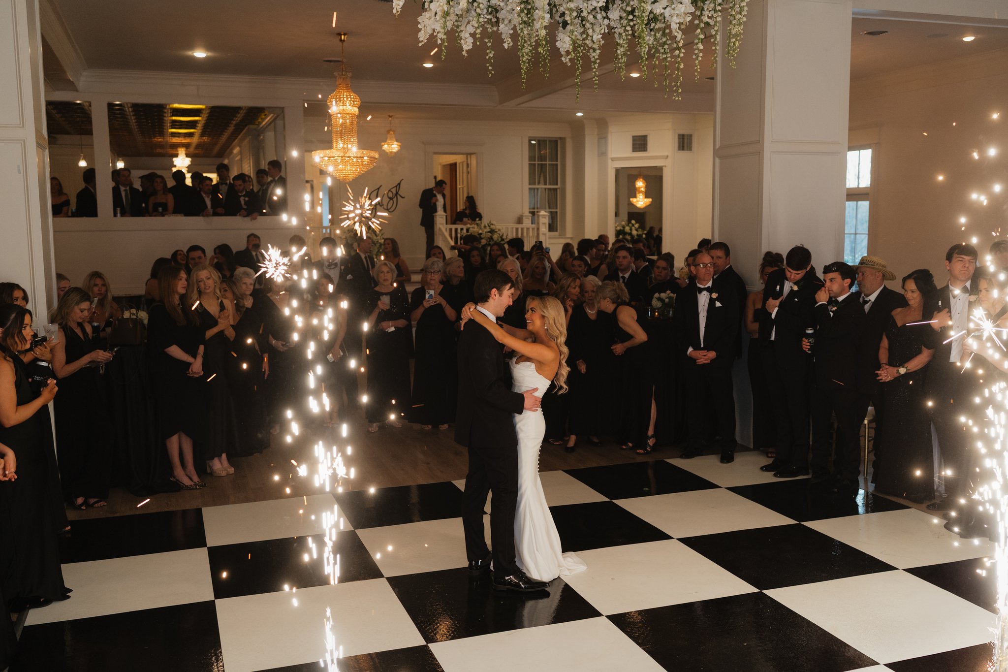 A wedding reception with a bride and groom dancing on a black and white checkered floor surrounded by guests, sparklers, large chandeliers, floral decorations, and string lights in Memphis, TN. 