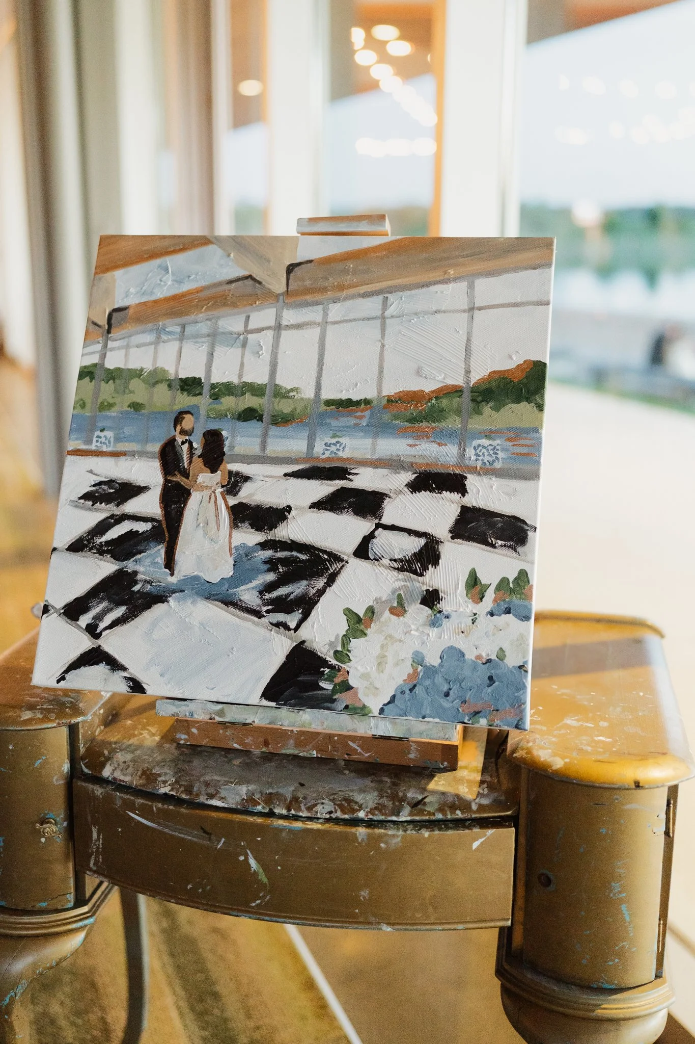 A small, painted canvas depicting a dance couple in front of large glass windows with a scenic outdoor view, on an easel inside a room with large windows in Memphis, TN. 