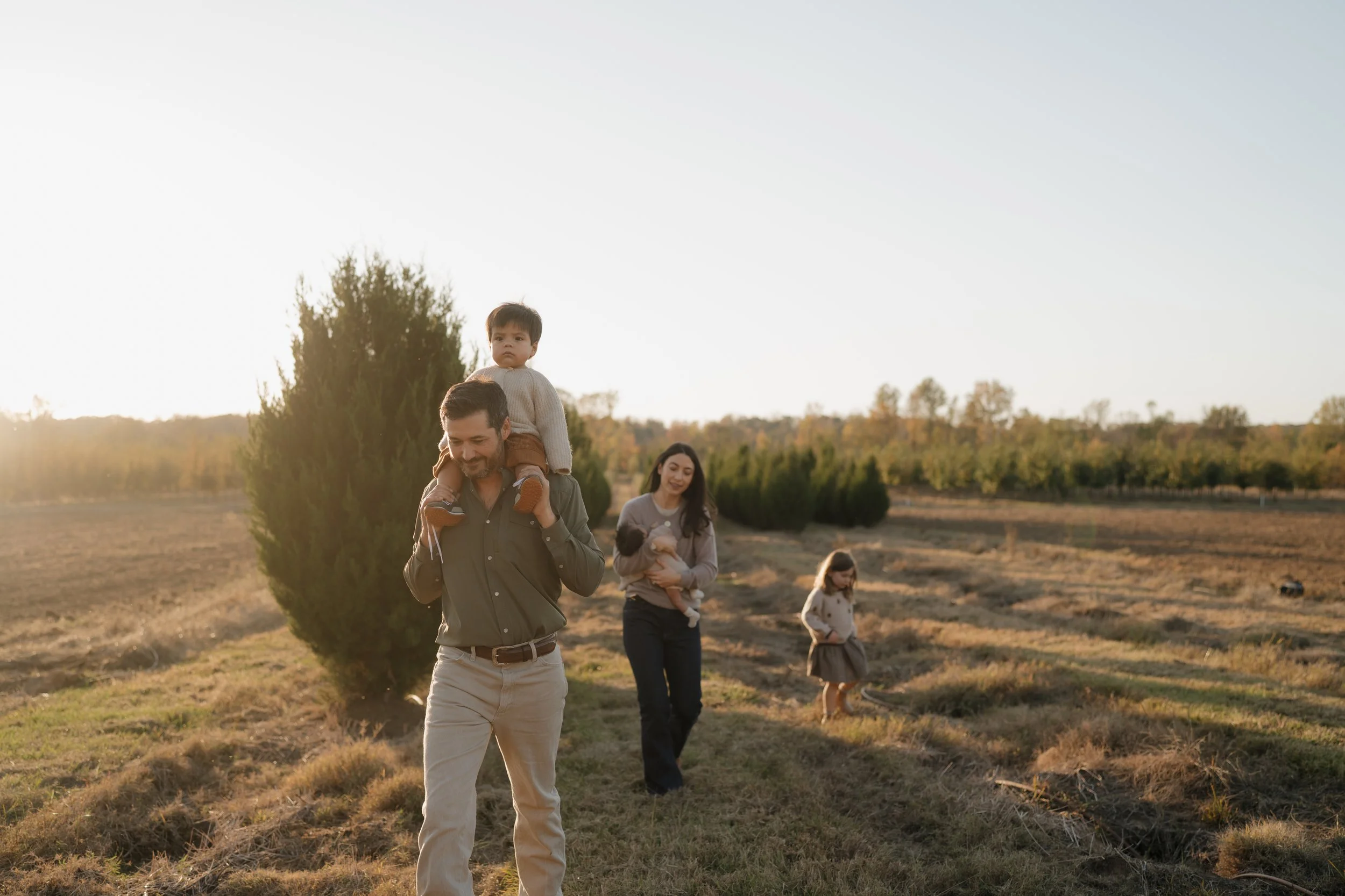 A family walking outdoors in a grassy field during late afternoon or early evening, with a man carrying a young boy on his shoulders, and a woman holding a child, all surrounded by trees under a clear sky in Mississippi.