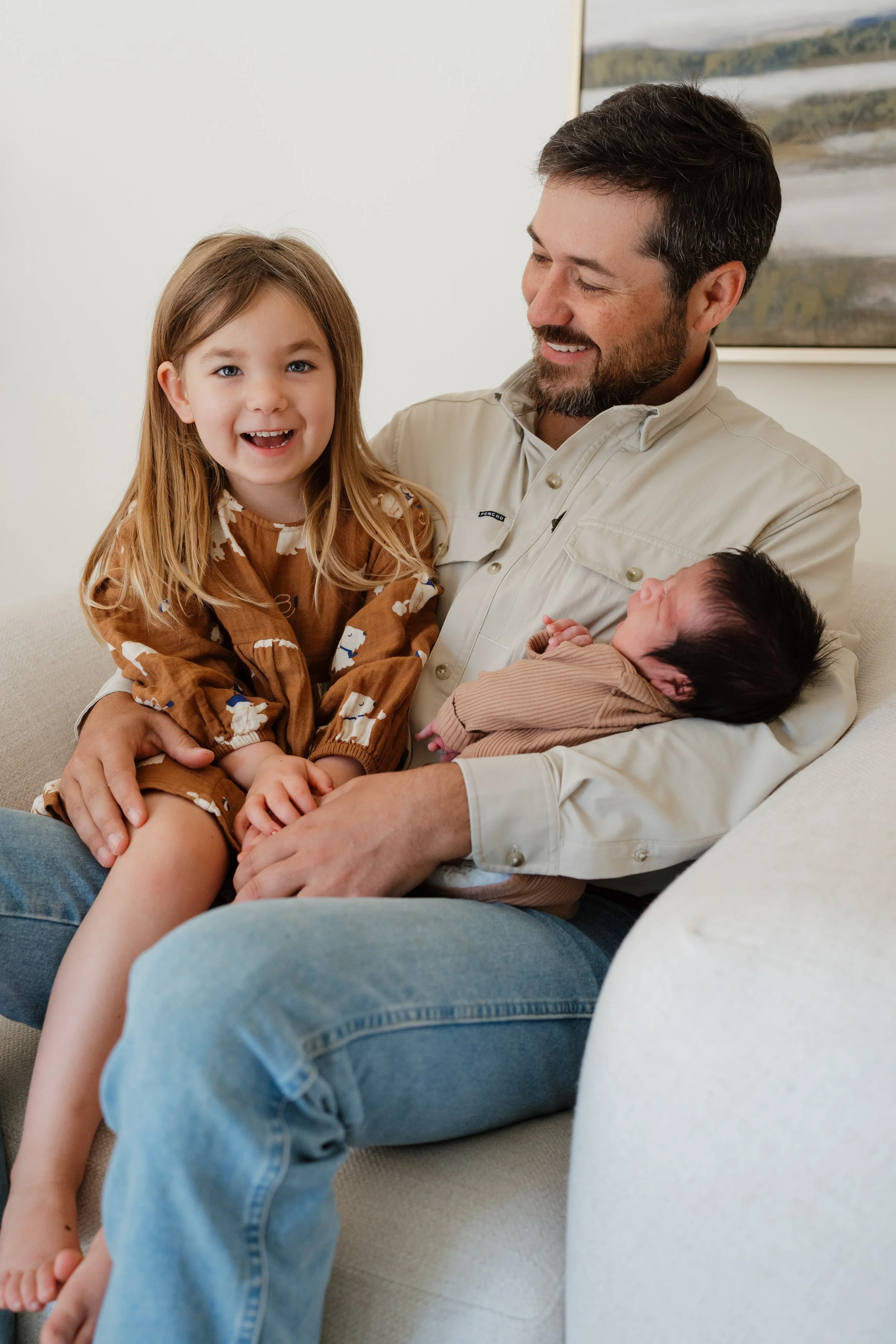 A man with a beard and short dark hair holding a newborn baby in his lap, with a young girl with long light brown hair and blue eyes sitting beside them, all smiling indoors in Memphis, TN. 