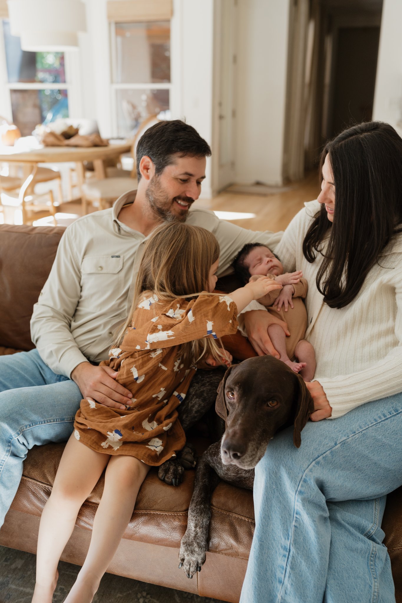 A family sitting on a couch with a dog, a toddler, and a newborn baby. The toddler is touching the baby's face, and the parents are smiling while looking at the baby. The dog is lying under the couch, with one paw on the baby's leg.