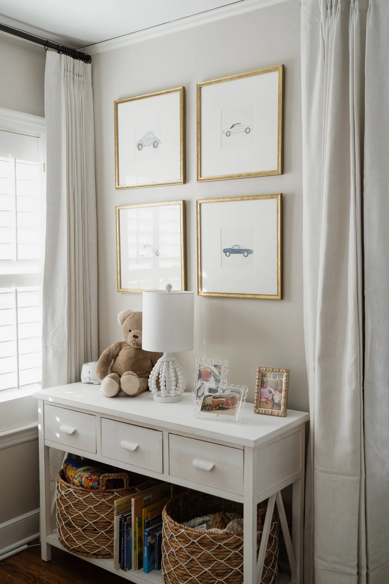 A white dresser with four drawers, topped with a teddy bear, a white lamp, framed photos, and a white decorative object. Two baskets with books and toys are on the lower shelf. The wall behind features four framed sketches of cars, surrounded by whit