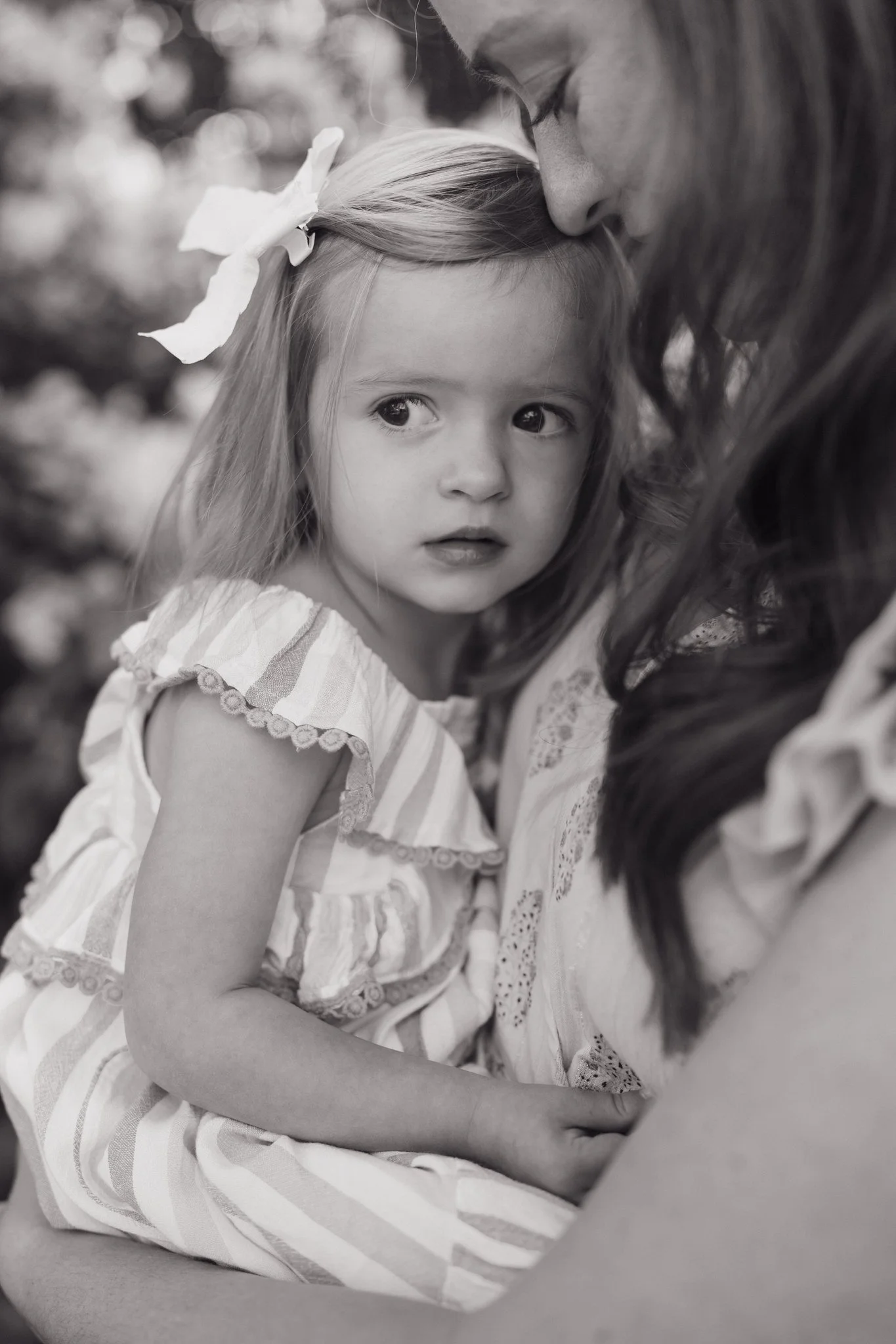 A black and white photo of a woman and a young girl in an outdoor setting. The girl, with long hair and a large bow on her head, is looking over her shoulder with a serious expression, while the woman is resting her forehead against the girl's head. 